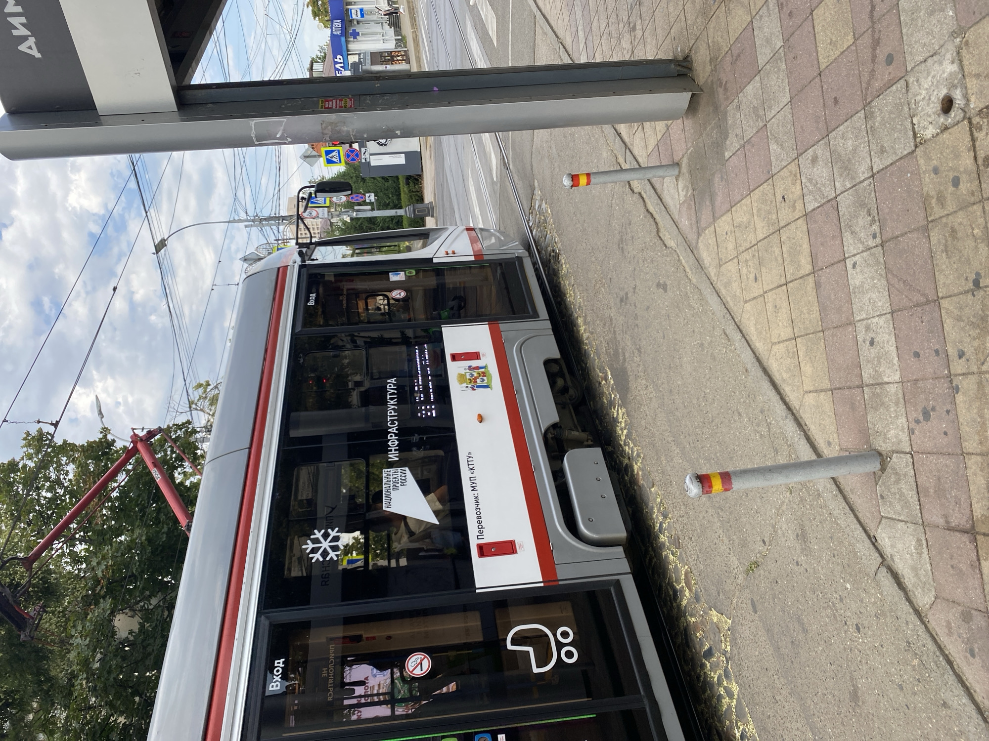 The image captures a section of a modern tram in the city of Krasnodar, Russia. The tram is gray and red with visible advertisements and signage on its windows. One sign displays a snowflake symbol and the text "НАЦИОНАЛЬНЫЕ ПРОЕКТЫ РОССИИ" (National Projects of Russia). Another sign reads "ИНФРАСТРУКТУРА" (Infrastructure). A smaller sign indicates "Перевозчик: МУП «КТТУ»" (Carrier: MUP "KTTU"), which is the municipal transport enterprise of Krasnodar. A symbol of a stroller suggests accessibility features on the tram.
The tram is positioned on tracks at what appears to be a street-level stop or crossing. In the background, there is a bus stop shelter with advertisements, and beyond that, buildings and trees. The sky is overcast with clouds. The foreground shows a sidewalk paved with square tiles in various shades of beige and pink, and a concrete curb. A gray metal bollard with red and yellow reflective bands stands on the sidewalk. The general scene depicts a typical urban transportation environment on a cloudy day.