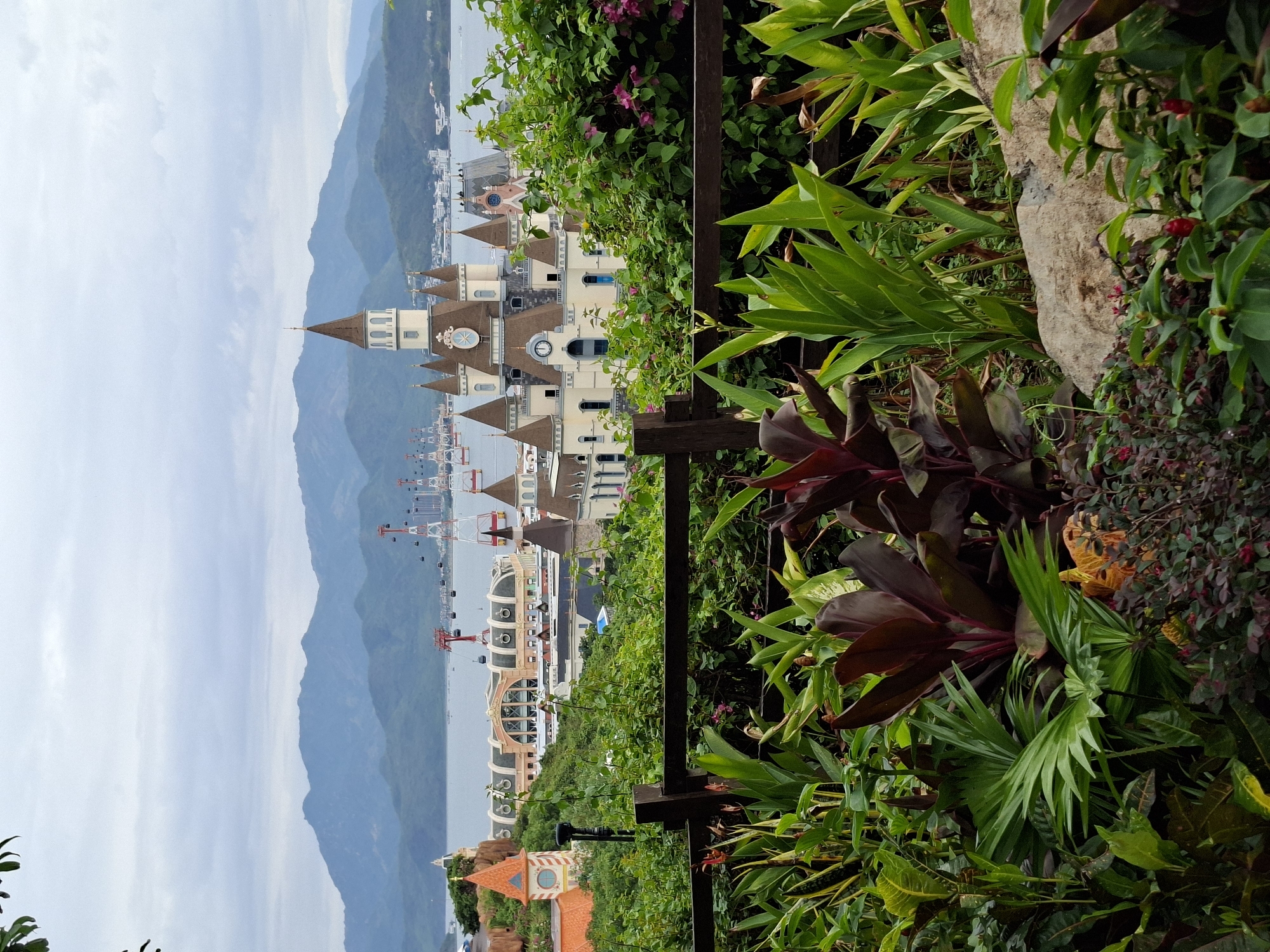 The image shows a view from a garden-like area with lush green foliage, red and green plants, and a wooden fence in the foreground. Behind the fence is a view of structures resembling European-style castles or buildings with towers and spires. In the background, there is a body of water with cable cars running above it, and beyond that, a mountain range under a slightly cloudy sky. The scene appears to be in a hilly or elevated area, overlooking the buildings and the cable car system. A section of Nha Trang Ward is visible in the background, as are sections of the city.