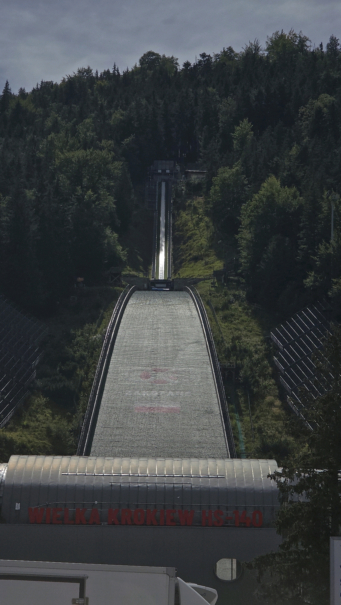 The image shows the Wielka Krokiew ski jumping hill, located in Zakopane, Poland. The ski jumping hill is a large, imposing structure made of concrete and metal, with a long, steep inrun ramp that leads to a curved outrun. The hill is surrounded by dense forest, with trees covering the slopes on either side. The sky is overcast, with dark clouds threatening rain.
The ski jumping hill is a prominent landmark in Zakopane, and it is a popular tourist attraction. The hill is used for professional ski jumping competitions, and it has also been used for other events, such as concerts and festivals. The hill is a symbol of Zakopane, and it is a reminder of the city's rich sporting heritage.
The image is a vertical shot, taken from a slightly elevated position. The ski jumping hill is the main subject of the image, and it is in focus. The trees and the sky are also visible in the background, but they are out of focus. The image is well-lit, and the colors are vibrant. The image is a good example of landscape photography, and it captures the beauty of the ski jumping hill and its surroundings.