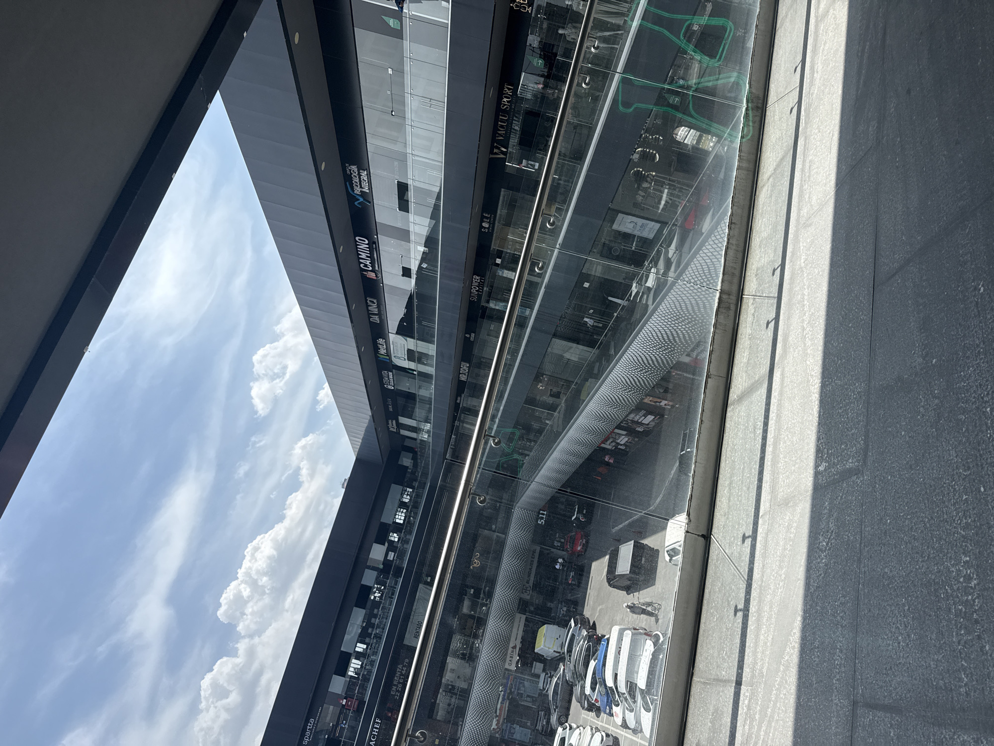 The image presents an upward-angled view of a multi-story commercial building, presumably a mall or shopping center, in San Bernardino Tlaxcalancingo, Mexico. The building features a modern architectural style with extensive use of glass, creating reflections of the sky and clouds. Various store names and logos are visible on the building's facade, including "spartta," "ACHEF," "MetLife," "DA VINCI," "CAMINO," "SUNPOWER," and "VACUU SPORT." Parked vehicles, mostly white, are visible at the base of the building, near the sidewalk. The edge of the image features a portion of the building's structure and the sky. The image is shot from a low angle, emphasizing the height of the building and the sky above.
