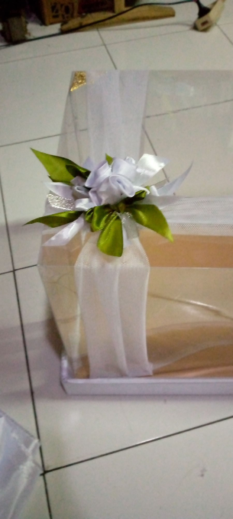 In Sumberwono, Indonesia, a white gift box is adorned with a satin ribbon flower and green satin leaves, draped with sheer white fabric. The box sits on a light-colored tiled floor. A gold fleck is visible on the upper left corner of the white fabric. The background is blurred, showing what appears to be a wooden object and electrical cords.