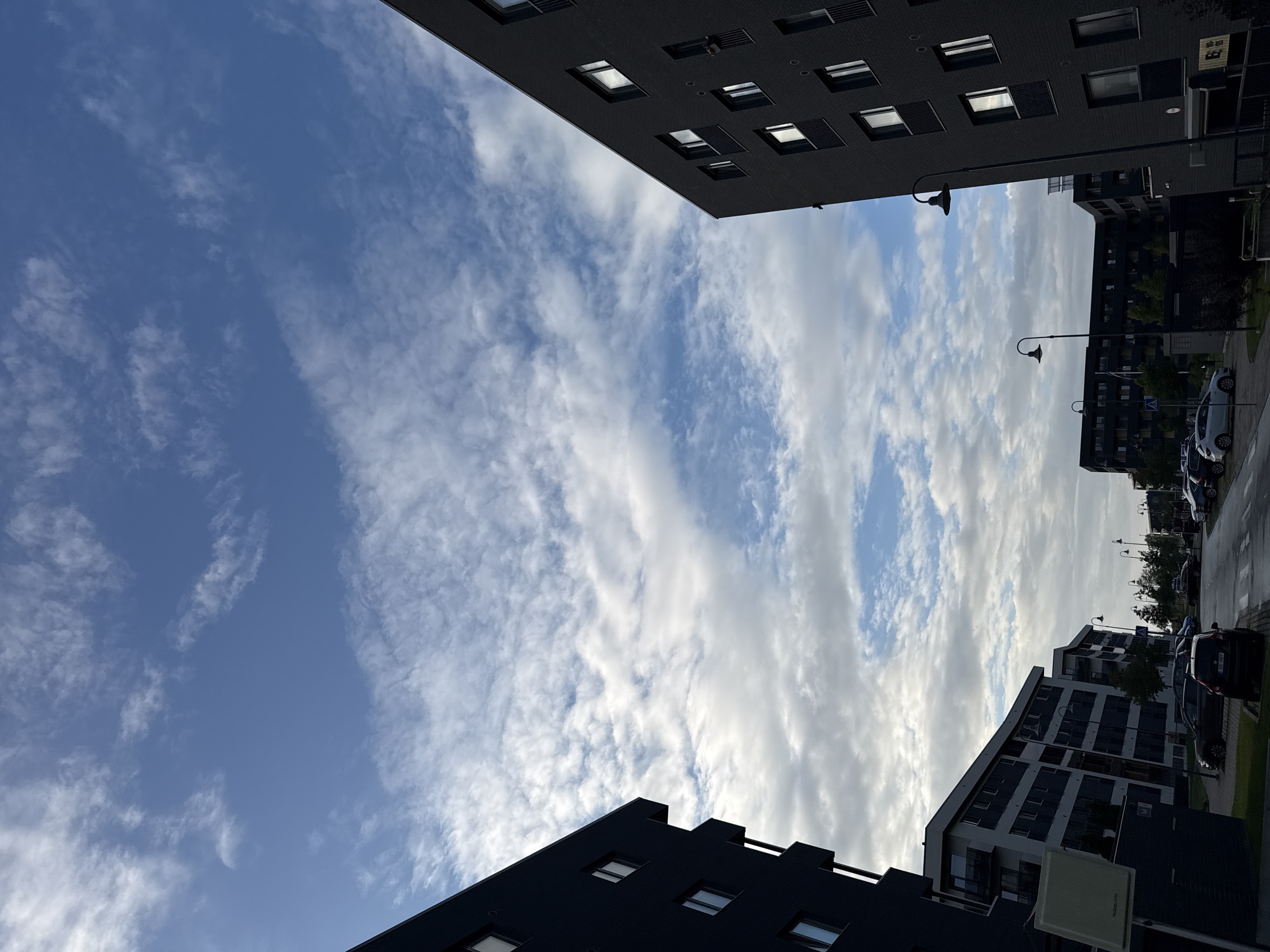 The image shows a street scene in Tallinn, Estonia. On the left, a dark blue apartment building occupies the foreground, angled towards the sky. To the right, a dark grey apartment building, also angled, rises into the frame. Between these two buildings, a street runs away from the viewer, lined with more apartment buildings in varying shades of grey and white. Cars are parked along the street and on the side. The sky is a mix of blue and white clouds. A street lamp is visible in front of the dark grey building on the right.