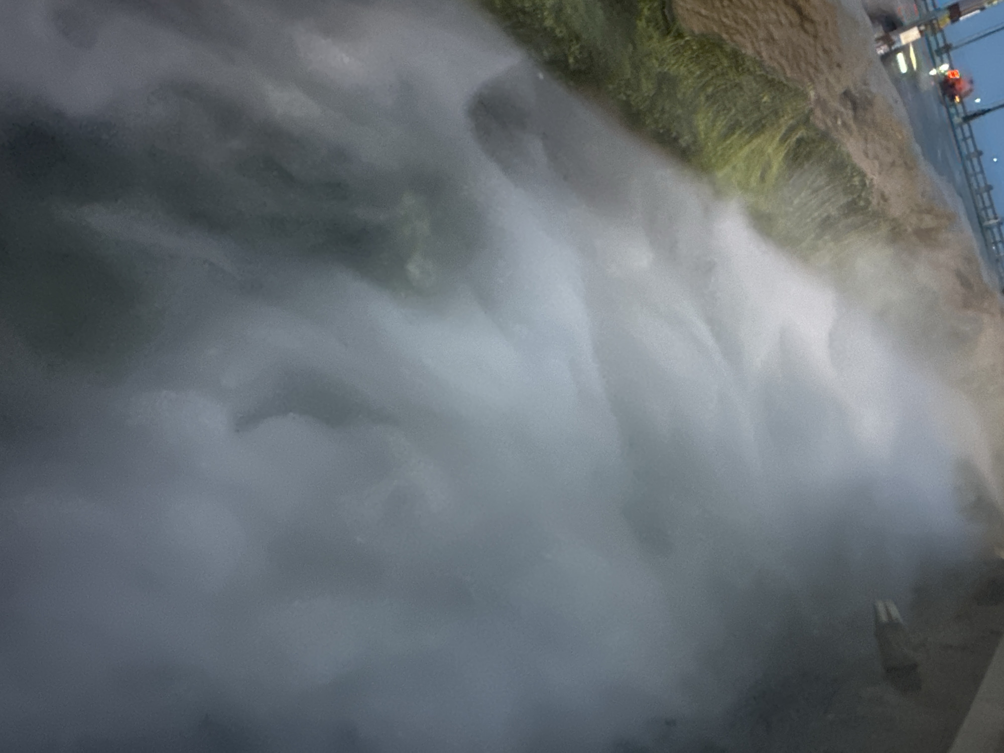 The image shows a waterfall in the city of Bac Nha Trang Ward, Vietnam. The water is rushing down, creating a mist that obscures much of the surrounding area. Vegetation is visible on the side of the waterfall, and a road with lights can be seen in the background.
