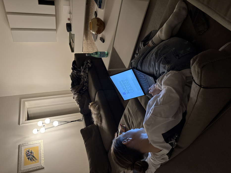 The image depicts a dimly lit domestic living room in Venice, Italy. A female individual, with hair pulled back, reclines on a dark grey sectional sofa, wearing a white long-sleeved shirt, blue jeans, and white socks. She is actively engaged with a black laptop resting on her lap, its illuminated screen displaying text and what appears to be a spreadsheet. Adjacent to her, on a different section of the sofa, a small, light-colored fluffy dog is asleep. A white rectangular coffee table in the foreground holds a green glass bottle of San Pellegrino, a brown and orange decorative vase, and two black remote controls. A black smartphone is visible on the sofa cushion near the individual's legs. The background features a tall black floor lamp with four spherical white lights, casting illumination, and a silver-framed artwork with a yellow background hangs on a light-colored wall. An open doorway leads to another room. Further right on the sofa, a pile of clothing or blankets is present, beneath a white wall-mounted cabinet system.