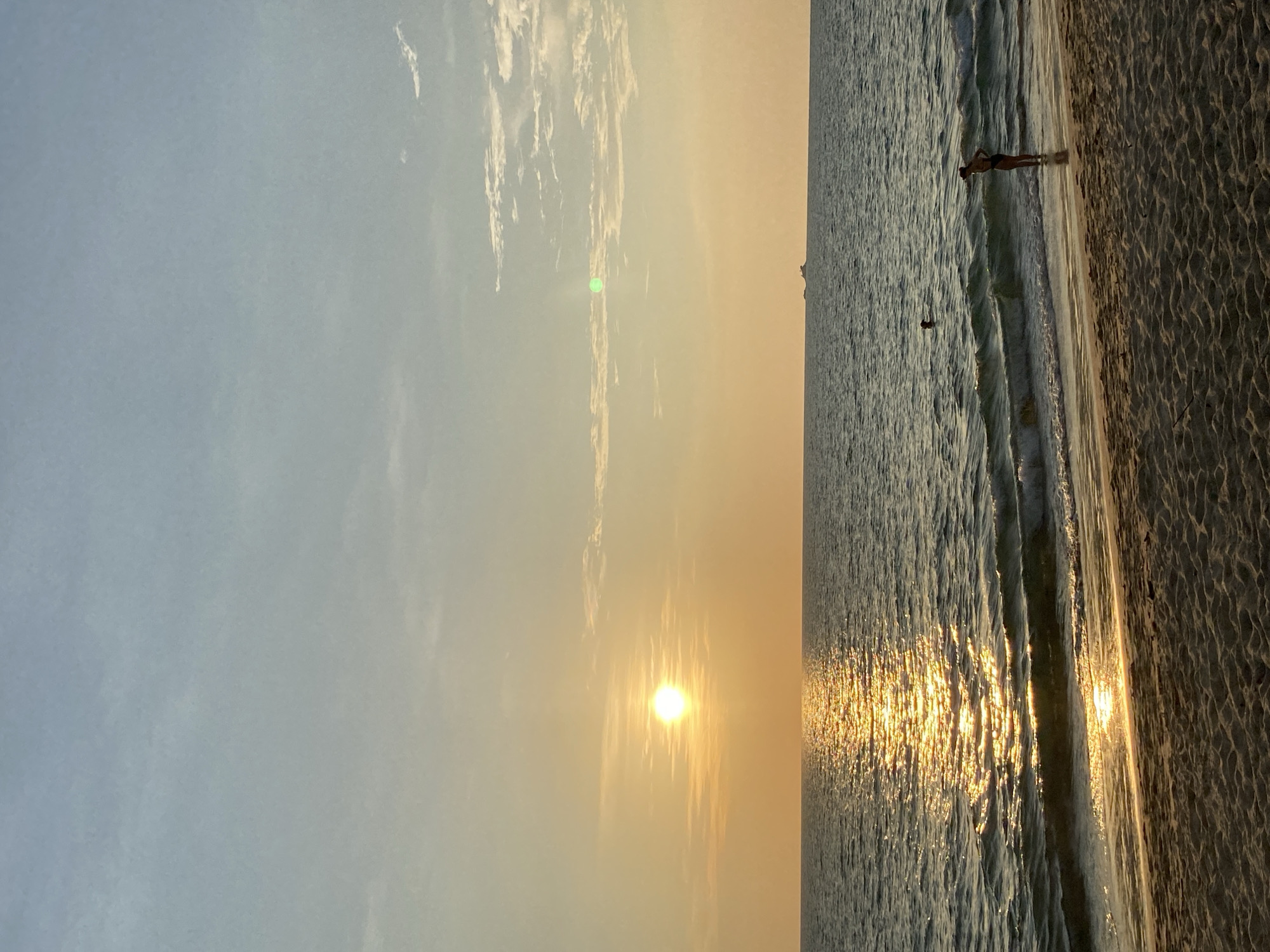 Captured at sunset in Choeng Thale, Thailand, the photograph displays a beach scene where the sun's golden light reflects vividly on the water's surface. A lone individual stands along the shoreline as the waves meet the sand, and another person is seen further out in the water. The sky is a gradient of light blue to warm yellow as the sun descends. The sand of the beach is visible on the right side of the image.