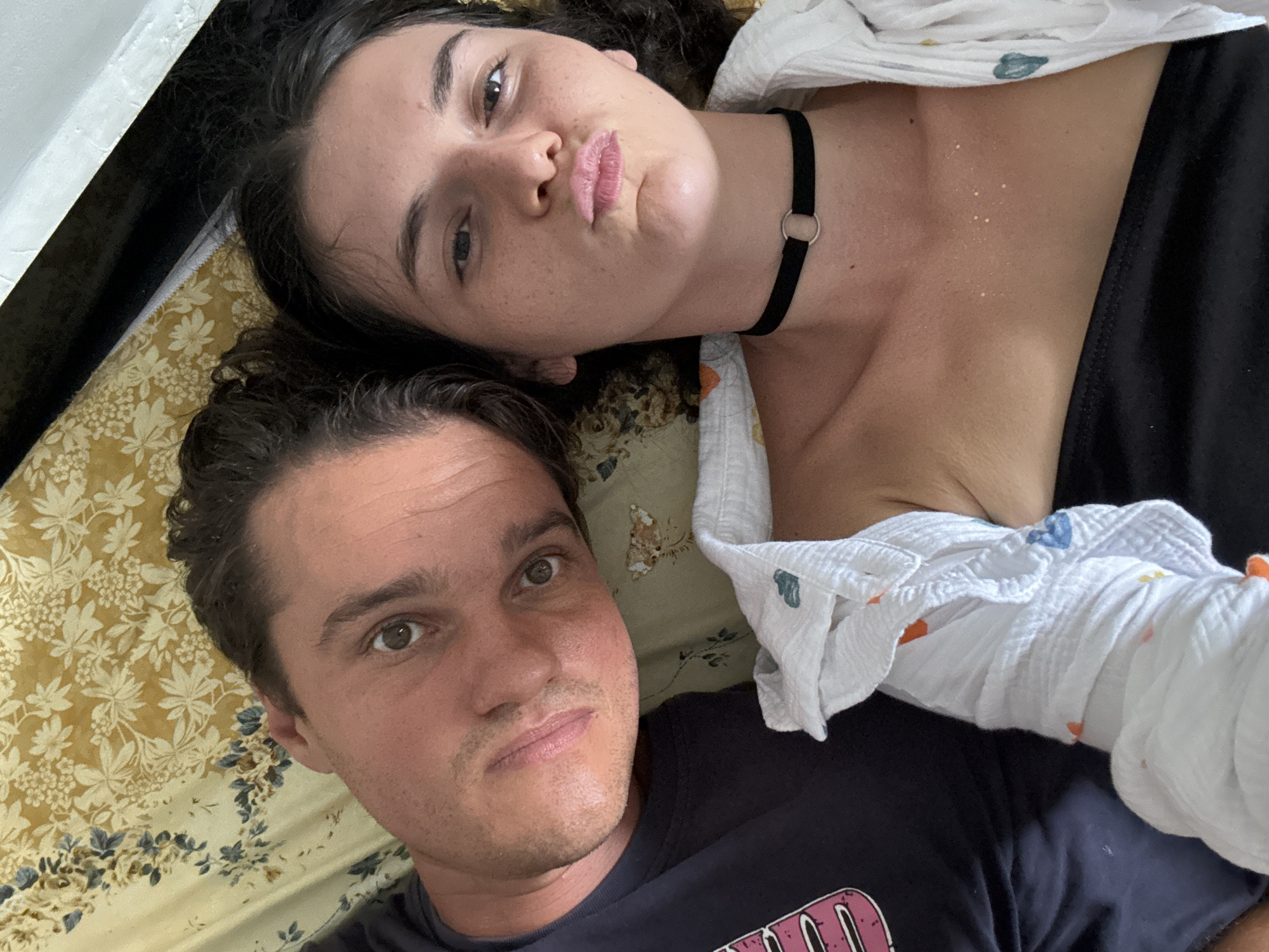 A selfie captured in an interior setting, likely a room, features two individuals lying on what appears to be a bed covered with a patterned sheet in yellow hues. The woman on the upper portion of the frame wears a black top, a black choker necklace, and has fair skin, dark hair, and is pouting. The man below her wears a dark blue t-shirt. A white cloth with multicolored designs rests partially on both individuals. The setting is illuminated by natural light, and a partial view of a white-painted structure is visible in the upper left corner of the frame. There is no immediate context to confirm the specific location in Mũi Né, Vietnam.