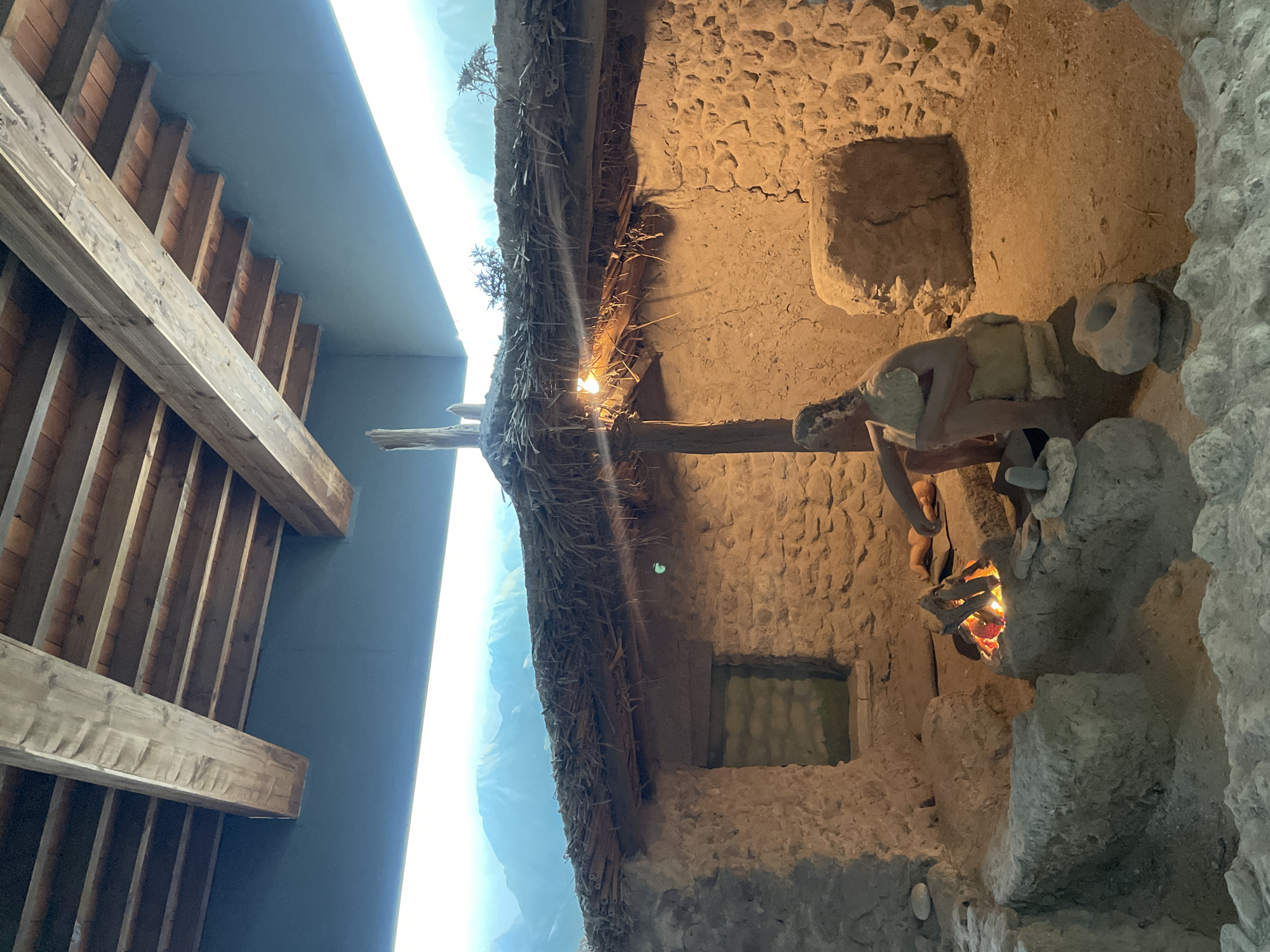 The image depicts a diorama of a prehistoric dwelling, possibly representing life in the vicinity of Kyrenia, Northern Cyprus. The dwelling is constructed with rough stone walls and a thatched roof supported by wooden beams. Inside, a figure, likely a mannequin, is seated next to a small, simulated campfire, appearing to be engaged in some activity. The environment is dimly lit, with the fire providing a warm glow. In the background, a painted backdrop suggests a mountainous landscape. The overall scene evokes a sense of ancient human settlement and daily life.