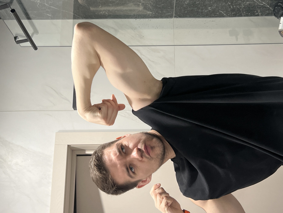 This image depicts a single male individual posing indoors, within what appears to be a residential bathroom in Kazan, Russia. The man, exhibiting short brown hair and light facial stubble, wears a black sleeveless top. An orange wristband is visible on his left wrist. He is actively flexing both biceps; his right arm (viewer's left) and left arm (viewer's right) are raised, elbows bent, with hands clenched into fists. His head is turned slightly to his right, and his gaze is directed in that direction, accompanied by a focused expression. The background features light-toned tiled walls, exhibiting a subtle veined pattern consistent with marble. A light-colored door frame is present on the left. To the right, a clear glass partition, likely a shower screen, is visible, equipped with a black, horizontally mounted towel bar or handle. A section of the wall beyond the glass partition displays darker, possibly black or dark grey, marble-patterned tiling. The lighting is uniform, suggesting an interior domestic setting.