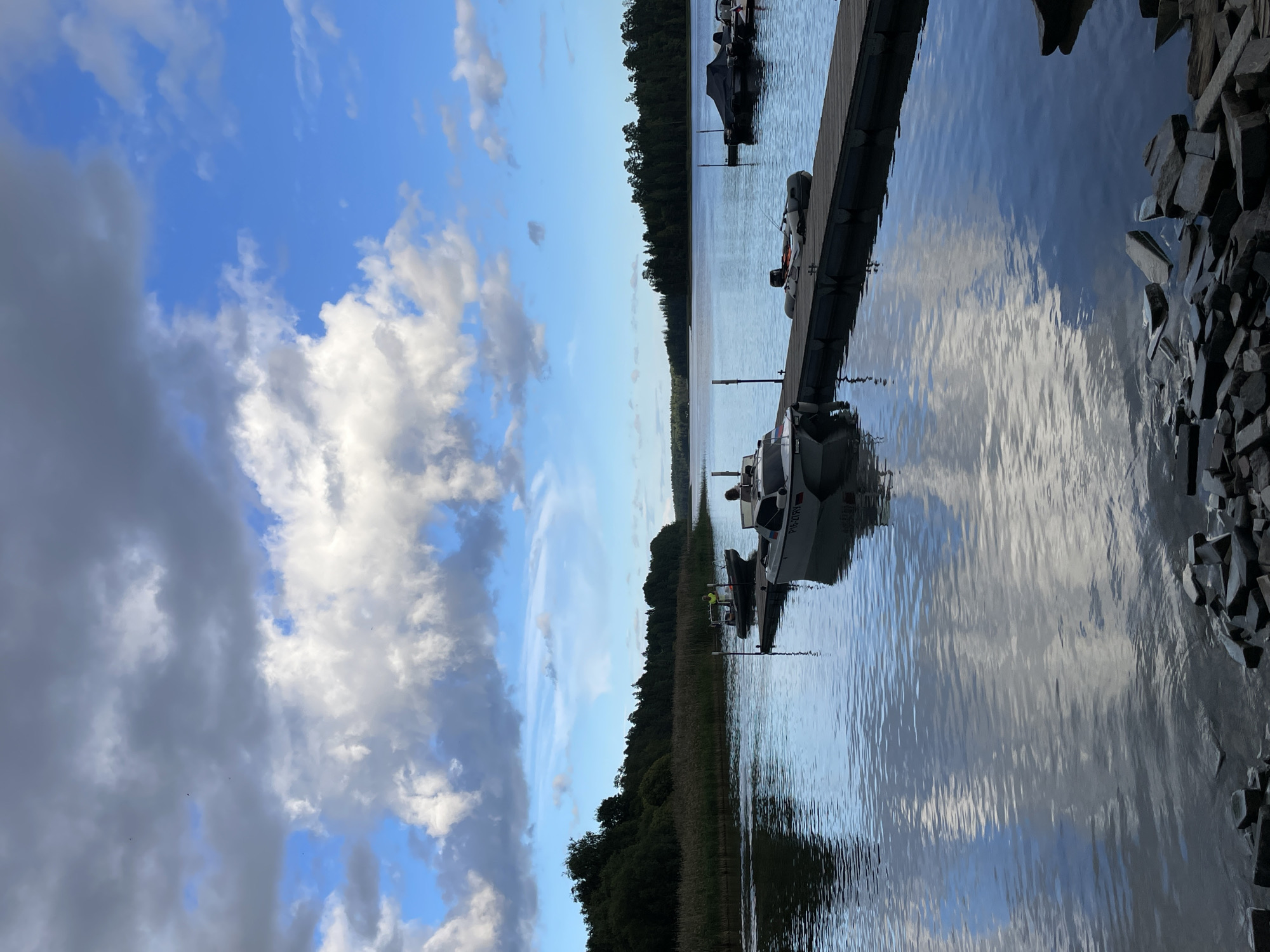 The image depicts a serene lake scene, possibly located in Berozovo, Russia, under a dramatic sky filled with a mix of dark and white clouds against a bright blue backdrop. A wooden dock extends into the calm water, where several boats are moored. One of these boats, a small white motorboat with the registration P16-27RY, is positioned at the end of the dock, facing away from the viewer. Another, slightly larger dark-colored boat with a person wearing a yellow shirt visible on board, is closer to the shore. A small inflatable dinghy with an outboard motor is also docked. The water reflects the sky and the surrounding vegetation, which includes reeds along the near shore and a dense line of trees forming the distant bank. The foreground shows a rocky shoreline. The overall atmosphere is tranquil, suggesting a quiet day at a lakeside location.