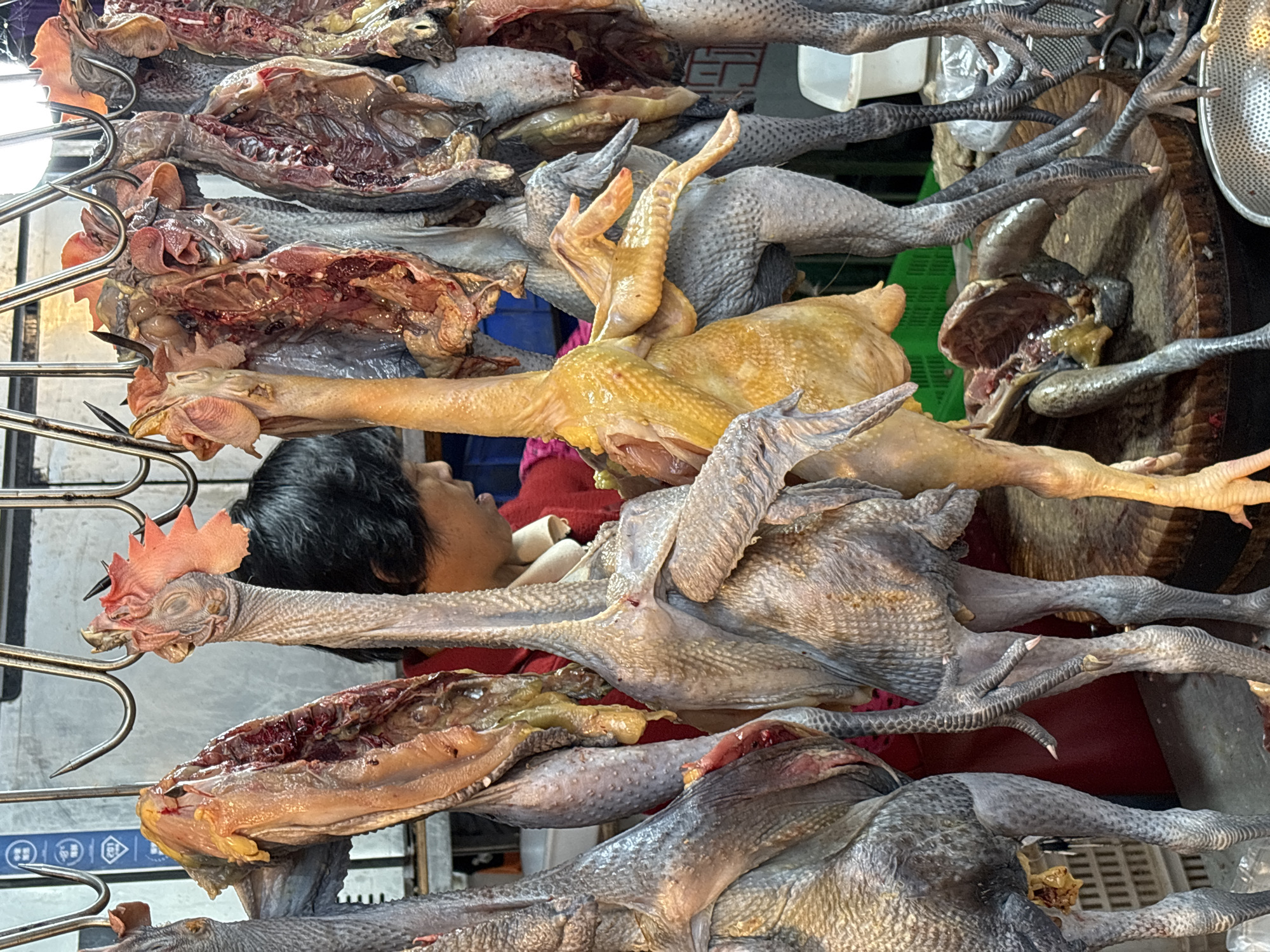 The image shows a street food scene in Jiangjin, China. There are several chickens hanging on metal hooks, some of which appear to be eviscerated. The chickens vary in color, with some being black-skinned and others yellow. A person with short, dark hair is partially visible behind the chickens. The background contains elements of a market setting, including a wooden cutting board and various containers. The scene appears to be a vendor selling poultry.