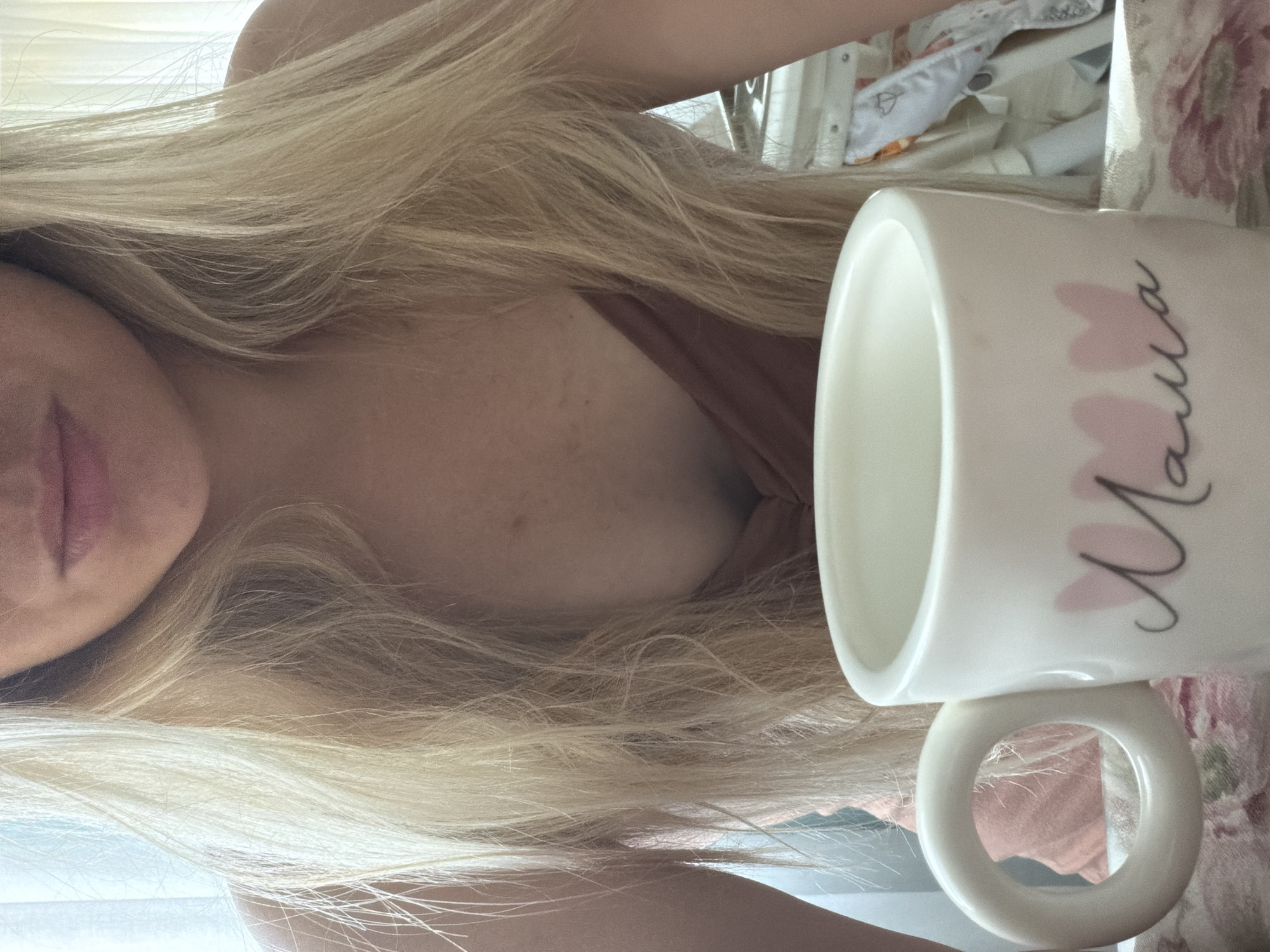 A close-up shot features a woman with long, blonde hair and fair skin. She is wearing a low-cut, pink top. In the foreground, a white mug with a distinctive rounded handle is visible. The mug is decorated with two pink heart shapes and the word "Mama" in cursive script. The background is softly blurred, with hints of white curtains and what appears to be a child's crib with patterned bedding. The lighting suggests an indoor setting, likely during the daytime. The image does not provide any discernible information about the city of Votkinsk, Russia.