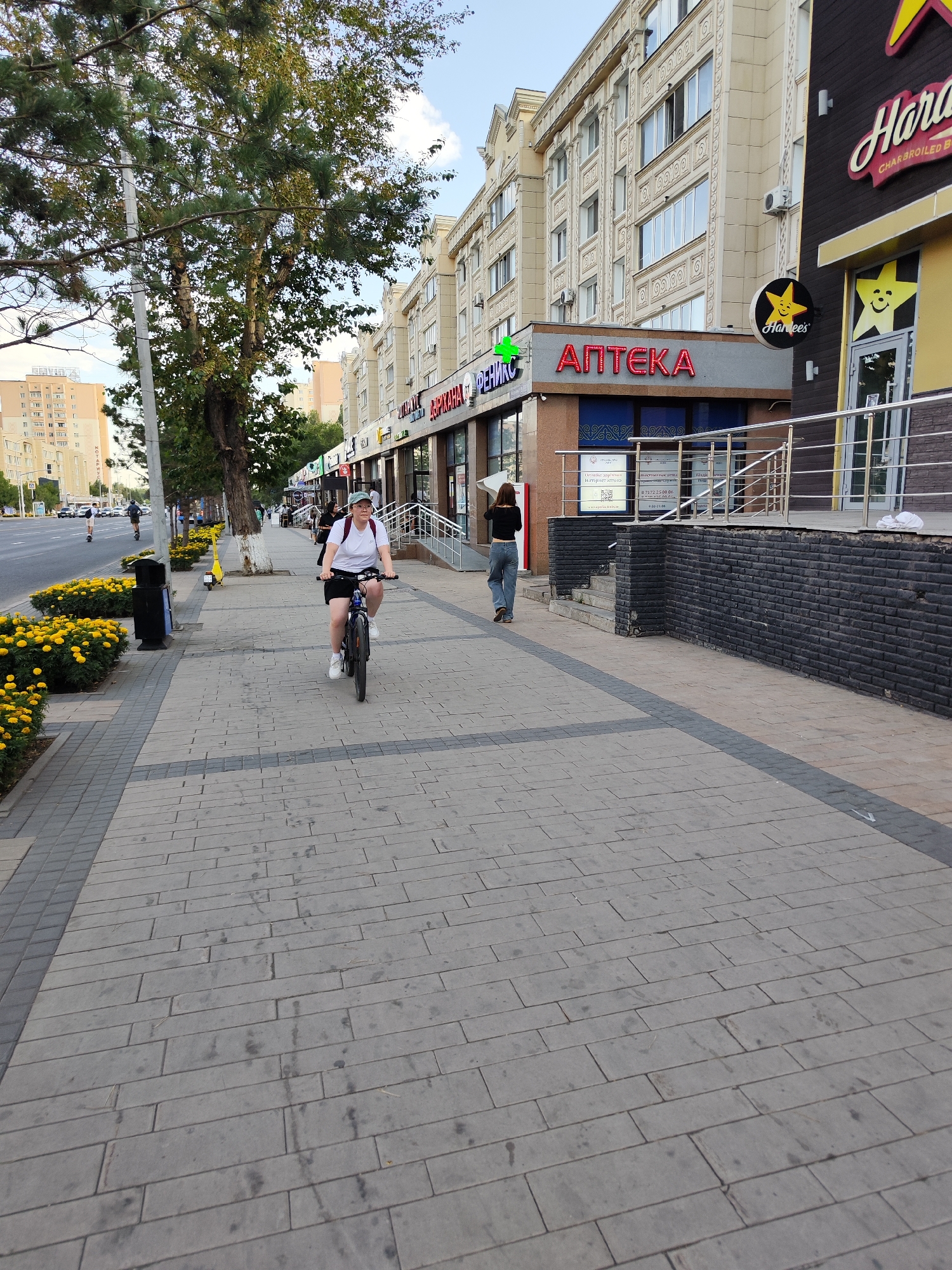A woman is riding a bicycle on a paved sidewalk in Astana, Kazakhstan. She is wearing a white t-shirt, black shorts, and a light green cap, and a backpack. The sidewalk is lined with yellow flowers and trees. To the right of the sidewalk is a multi-story building with several businesses, including a pharmacy with a green cross and red lettering that says "АПТЕКА" (Apteka), and a restaurant with a "Hardee's" sign. A woman in a black top and blue jeans is walking away from the camera on the sidewalk. The street is visible to the left of the sidewalk, with cars and other people. The sky is clear and blue.