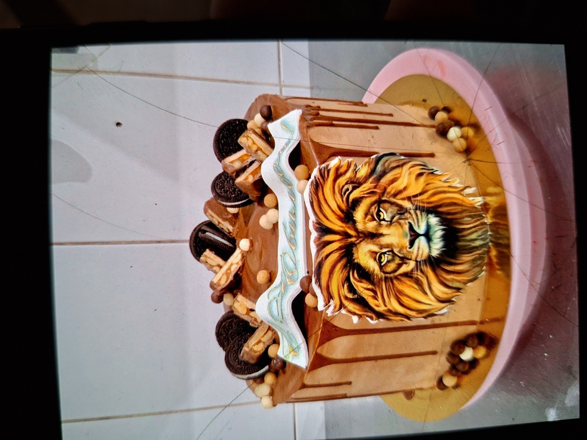 This media file shows a close-up, slightly tilted, top-down view of a decorative cake. The cake is covered in chocolate frosting and adorned with various toppings. Prominently featured on the side of the cake is an image of a lion's head with a golden mane. Around the cake, there are several chocolate sandwich cookies (like Oreos), pieces of candy bars, and small spherical candies in light brown and white colors. The cake rests on a pink and gold cake stand, which is on a white tiled surface. The lighting appears to be artificial, casting soft shadows. There is no discernible text in the image. The scene appears to be indoors, possibly in a home or a bakery, given the tiled surface. The overall presentation suggests it is a celebratory cake, possibly for a birthday or a special event. The image quality is slightly degraded, with visible lines and smudges on the screen capturing the image.