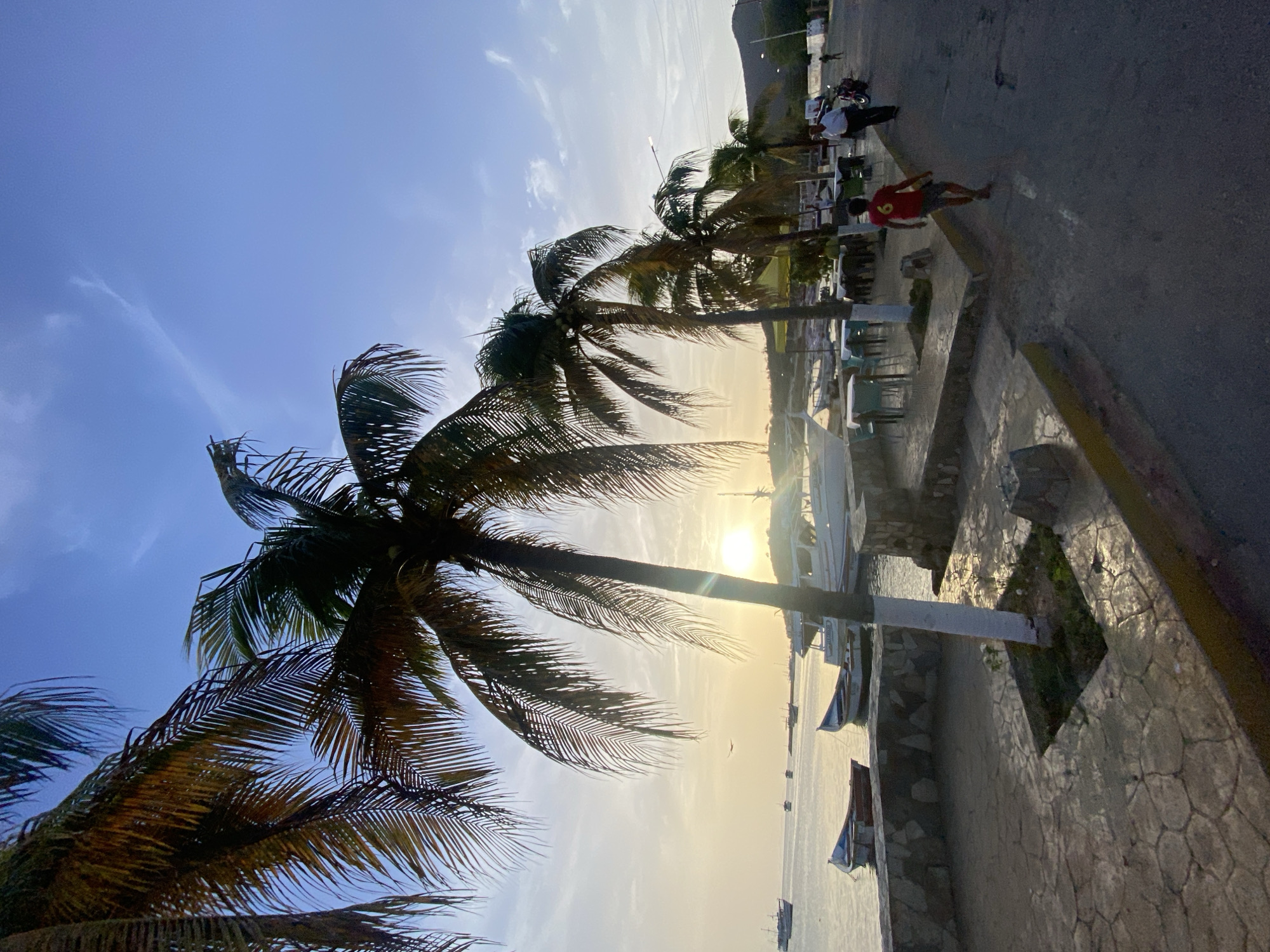 The image depicts a coastal scene in Juan Griego, Venezuela, during what appears to be late afternoon or early evening. The sun, low in the sky, casts a warm, golden light, creating a soft glow over the water and illuminating the clouds. A prominent line of palm trees with fronds spread against the blue sky stretches along the waterfront.
In the foreground, a paved walkway with irregularly shaped stones borders a stone wall overlooking the water. Several small boats are moored near the shore. Beyond the walkway, an outdoor seating area with tables and chairs is visible, suggesting a restaurant or cafe.
Further back, a paved road runs parallel to the waterfront. A young boy wearing a red shirt with the number six on it walks along this road, away from the camera. An adult man in a white shirt and dark pants walks in the same direction, with a motorcycle parked nearby. In the distance, more boats are anchored in the bay, and hills form the backdrop against the horizon. The overall atmosphere is serene and picturesque, characteristic of a tropical coastal town.