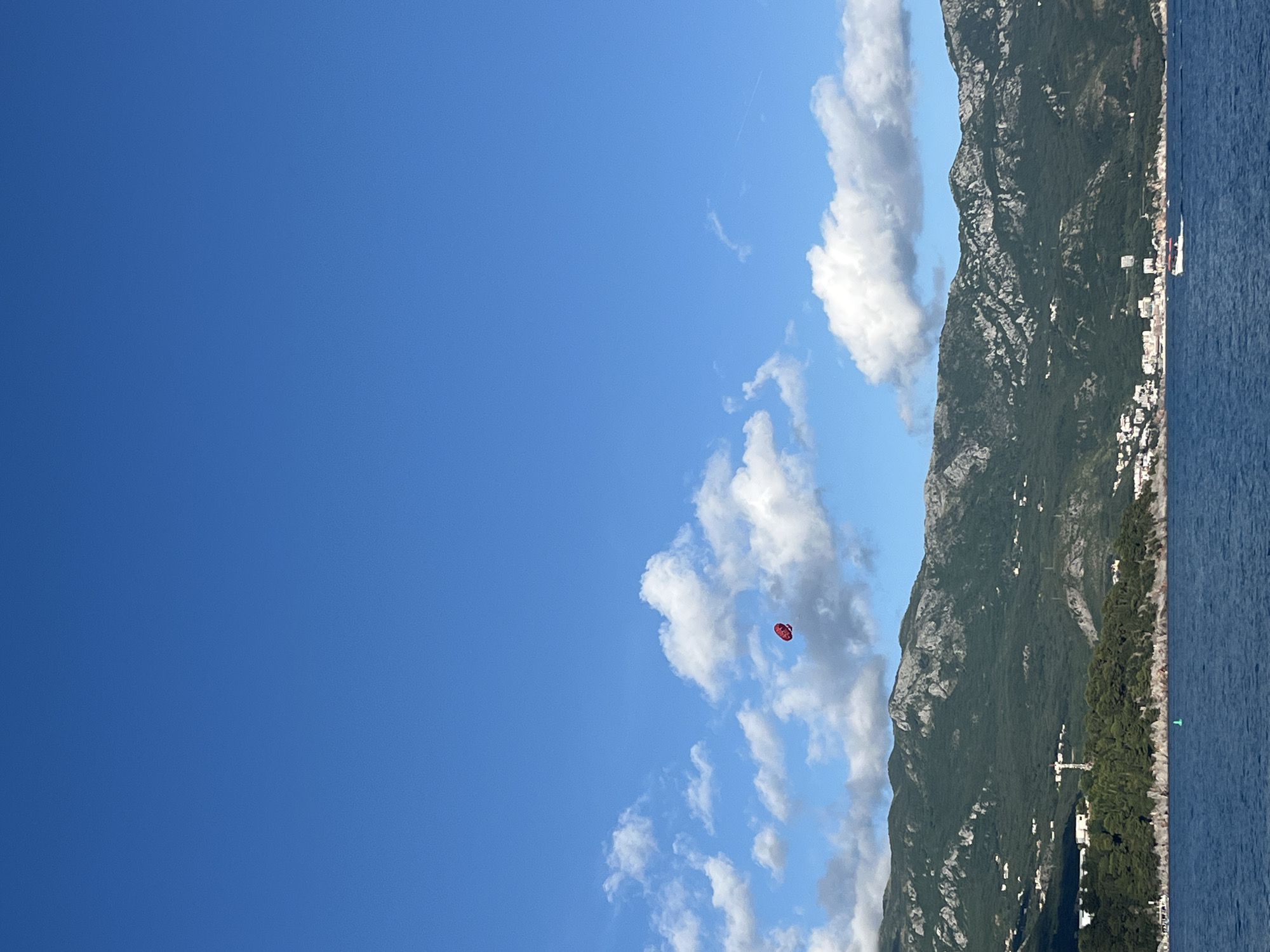 This vivid landscape image captures a scenic coastal view, likely around Budva, Montenegro, under a bright, clear blue sky dotted with fluffy white clouds. The top two-thirds of the frame are dominated by the expansive sky, where a small, bright red parasail with a person attached is visible, suggesting a recreational water sport activity.
Below the sky, a majestic, rugged mountain range, lush with green vegetation and rocky outcrops, stretches across the middle and right side of the frame. At the base of the mountain, along the coastline, a settlement with numerous white buildings indicates a vibrant coastal town. The bottom third of the image features the deep blue sea, reflecting the clear sky. A small white boat can be seen cruising on the water in the distance, and a green buoy is positioned closer to the shore.
The scene conveys a sense of summer leisure and beautiful natural surroundings, typical of a sunny day with excellent weather for outdoor activities. There is no visible text in the image.