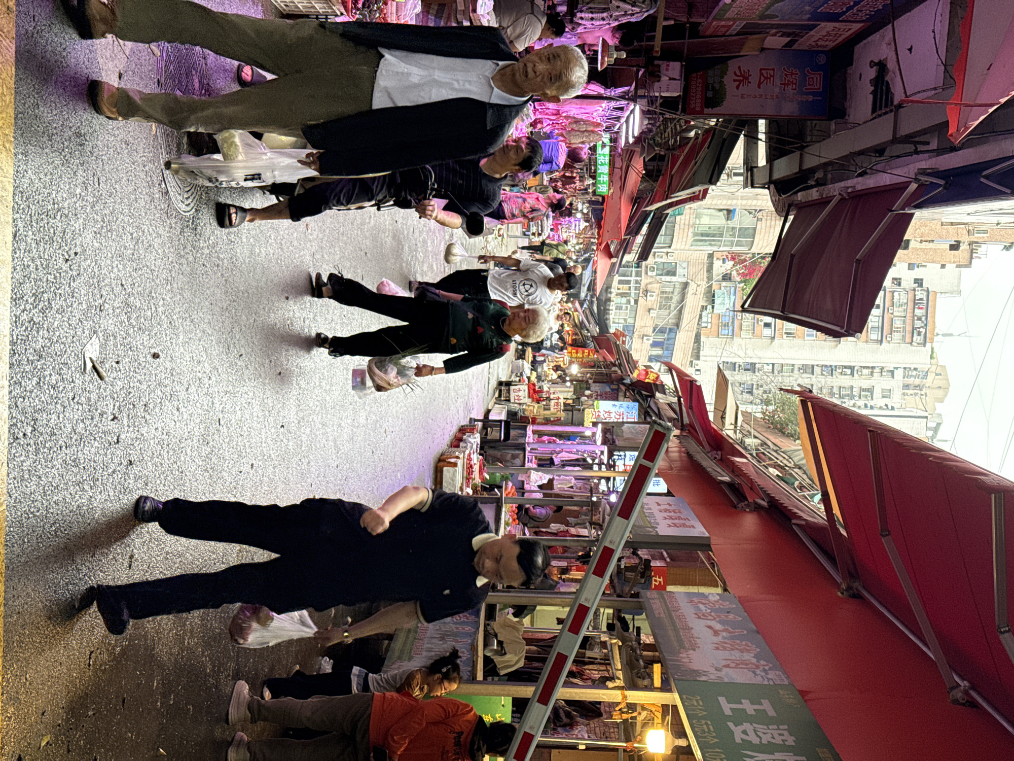 Here's a detailed description of the image:
**General Overview:** The image depicts a bustling street scene in what appears to be a market in an urban setting. The perspective is a street-level view, looking down a narrow lane filled with vendors, shoppers, and various commercial structures.
**Location:** The image is likely captured in Nan'an District, China based on OCR text.
**People:**
* Numerous individuals of various ages are present in the scene. They are mostly pedestrians moving through the lane.
* There are elderly people, younger adults, and children present.
* Some are dressed in casual clothing, while others wear more formal attire.
* A few are carrying shopping bags, suggesting they are customers.
**Objects:**
* A variety of goods are displayed in stalls lining the lane, indicating this is a marketplace.
* The presence of storefronts and overhead awnings suggests established businesses.
* There's a red and white traffic barrier partially blocking the lane.
* Signboards with Chinese characters are visible above the storefronts and awnings.
**Actions & Interactions:**
* People are walking, presumably shopping or passing through the market.
* Vendors are likely engaging with customers, although specific transactions aren't visible.
* The barrier suggests a level of traffic control or access restriction.
**Environment:**
* The lane is narrow and crowded, with pedestrians walking along its length.
* Buildings of varying heights line both sides of the street.
* Overhead awnings and signs create a visually cluttered environment.
* The lighting suggests it is either daytime or early evening.