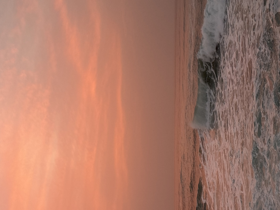 The scene displays an ocean at sunset. The sky is a soft gradient of orange and pink, with subtle cloud formations. The ocean reflects the sky's colors, creating a warm, golden tone across the water. White waves crash near the shore, forming a foamy texture. The image is vertically oriented, emphasizing the vastness of the sky and the movement of the sea.