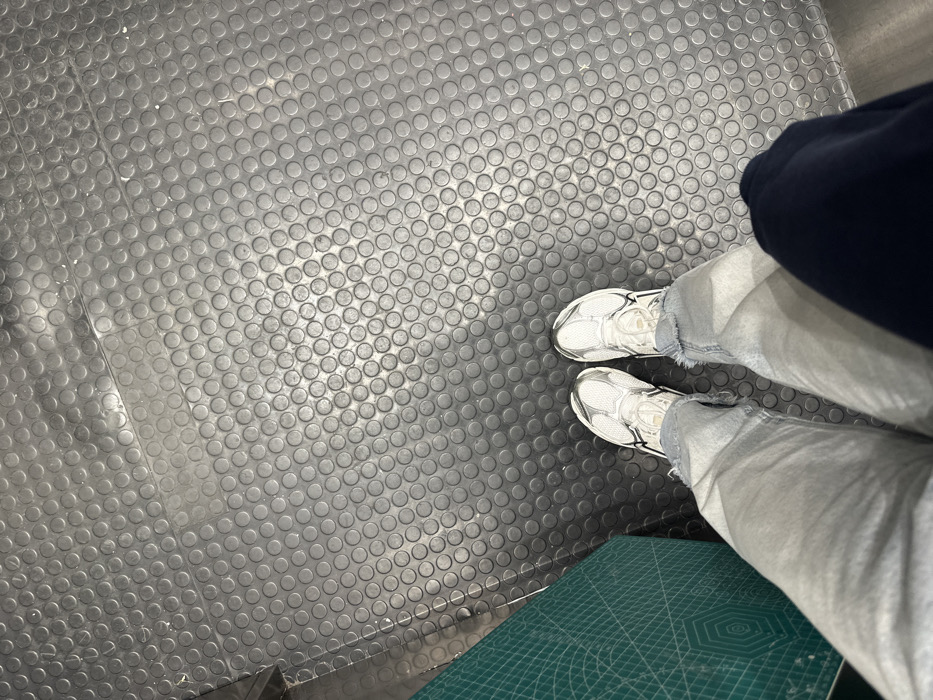 A high-angle shot depicts a person standing on a black rubberized floor with a repeating circular pattern. The individual wears white sneakers and light wash jeans with distressed hems. A portion of a dark blue garment, possibly a hoodie or jacket, is visible at the top right. A green cutting mat with geometric patterns lies at the bottom right corner, partially obscured. The lighting appears to be diffused, creating soft shadows across the scene.