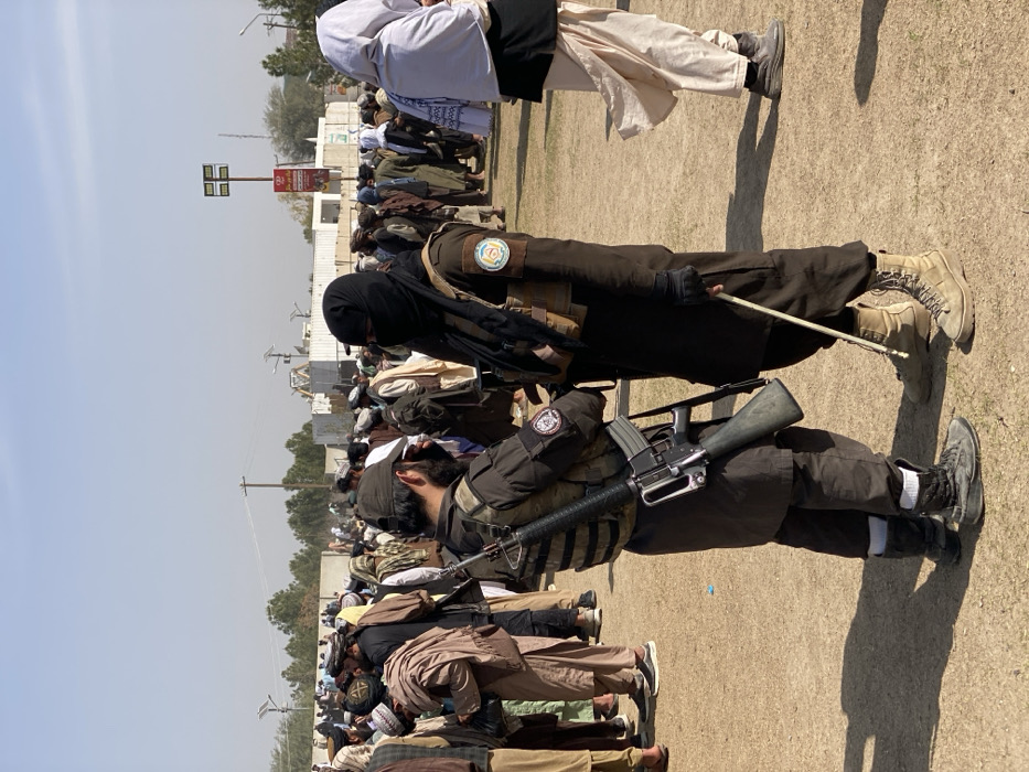 Men are praying in rows outdoors on a tan dirt surface. Many are wearing traditional clothing. At least two men are dressed in tactical gear with rifles slung across their chests. One has a black face covering, and the other has a patch on his shoulder. Some wear light-colored turbans or hats. A light post is visible in the background, along with buildings and trees.