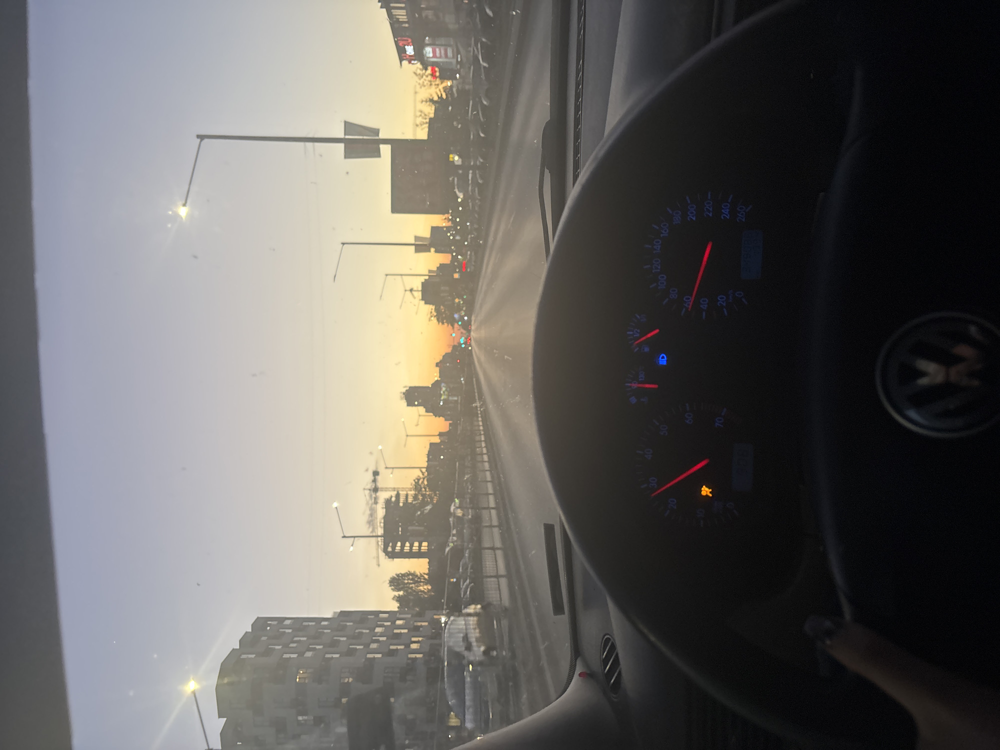 The image shows a view from inside a car, likely the driver's perspective. The dashboard and steering wheel of a Volkswagen are visible, with a speedometer reading approximately 80 km/h and a check engine light illuminated. Outside, a city street scene unfolds during what appears to be sunset or early morning. Buildings and streetlights line the road. Based on the layout and the presence of specific buildings (including some under construction), this scene is likely in Sofia, Bulgaria.