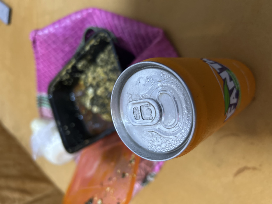 The scene, located in Ilishan, Nigeria, depicts an arrangement of food and beverage items on a light brown surface with faint, dark stains. In the foreground, a vertical, unopened orange Fanta soda can with significant condensation on its aluminum top and sides is positioned. Behind and to the right of the can, a black rectangular plastic container holds a dark, viscous substance intermixed with lighter, granular, possibly greenish particulate matter, suggesting food remnants. A bright pink woven fabric or cloth is partially visible underneath and around this container. To the left, a portion of a translucent orange plastic bag containing blurred dark specks is visible. Further back, a small, crumpled white plastic item is discernible. No individuals are present in the frame, and no active interactions are depicted.