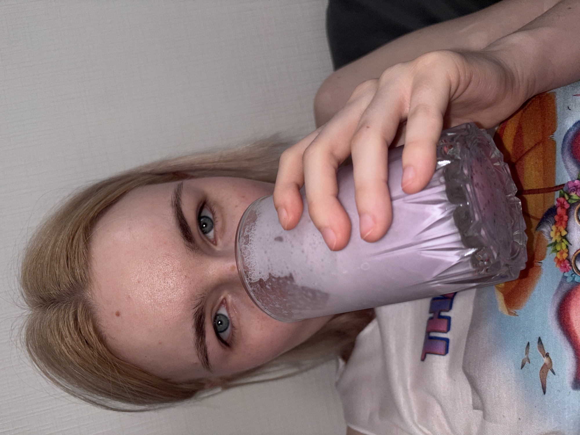 This image, taken in Sinyavino, Russia, features a fair-skinned individual with light blonde hair parted centrally and blue eyes, making direct eye contact with the camera. A small, dark mole is present on the individual's forehead, and faint freckles are visible on the upper cheeks. The subject wears a light-colored t-shirt displaying a colorful graphic design with partial letters "TH" and what appears to be a cartoon image incorporating birds and flowers. The individual's right hand holds a clear, faceted glass containing a light purple liquid topped with foam, obscuring the lower face. The background consists of a plain, light-colored textured wall, with a dark-colored object, likely furniture, partially visible on the right.