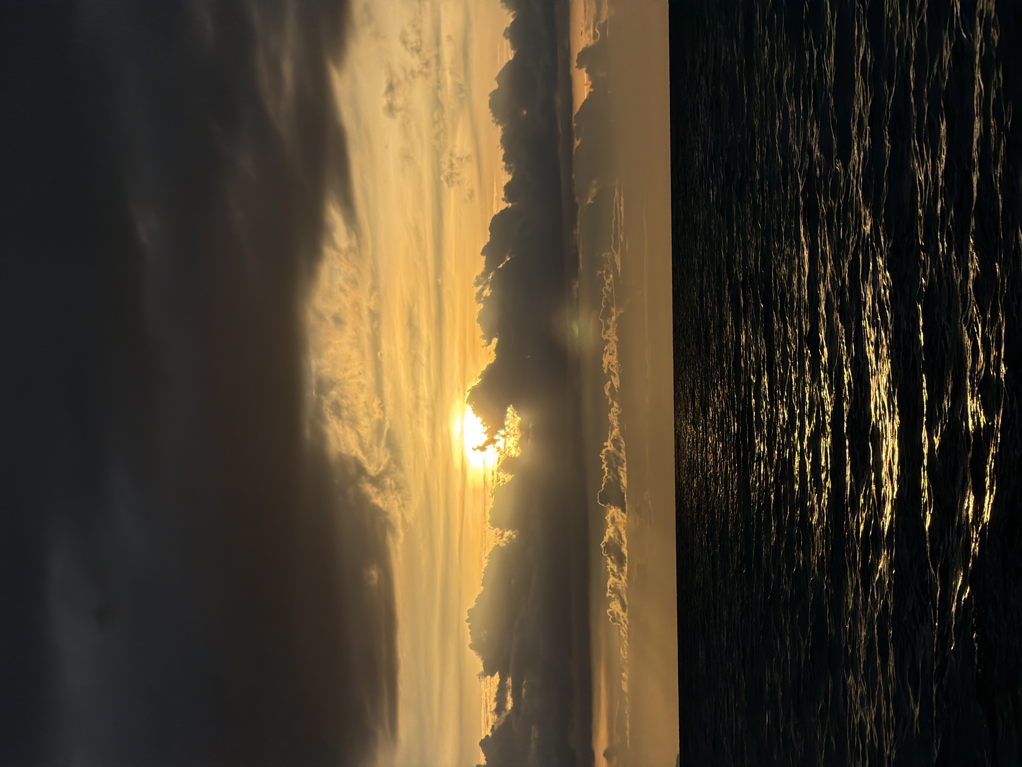 This media file depicts a serene sunset over the ocean at Baan Plai Laem, Thailand. The scene is divided into two distinct halves: the sky and the water.
The sky is filled with dramatic clouds, illuminated by the setting sun. The sun itself is partially obscured by dark, heavy clouds, allowing golden rays to pierce through and create a warm, ethereal glow. Lighter, wispy clouds stretch across the horizon, catching the light and adding depth to the sky. The overall impression of the sky is one of grandeur and natural beauty.
The lower half of the image is dominated by the dark, rippling surface of the ocean. The setting sun's light reflects off the waves, creating streaks of shimmering gold that stretch across the water. The contrast between the dark sea and the bright reflections is striking, conveying a sense of peaceful vastness.
The time of day is clearly sunset, given the warm, low-angled light and the dramatic cloud formations. The weather appears calm, with only gentle ripples on the water. The location is identified as Baan Plai Laem, Thailand, which suggests a tropical or subtropical setting. There are no people or objects visible in the frame, focusing solely on the natural landscape. The overall emotion conveyed is one of tranquility and awe at the beauty of nature. No text is visible in the image.