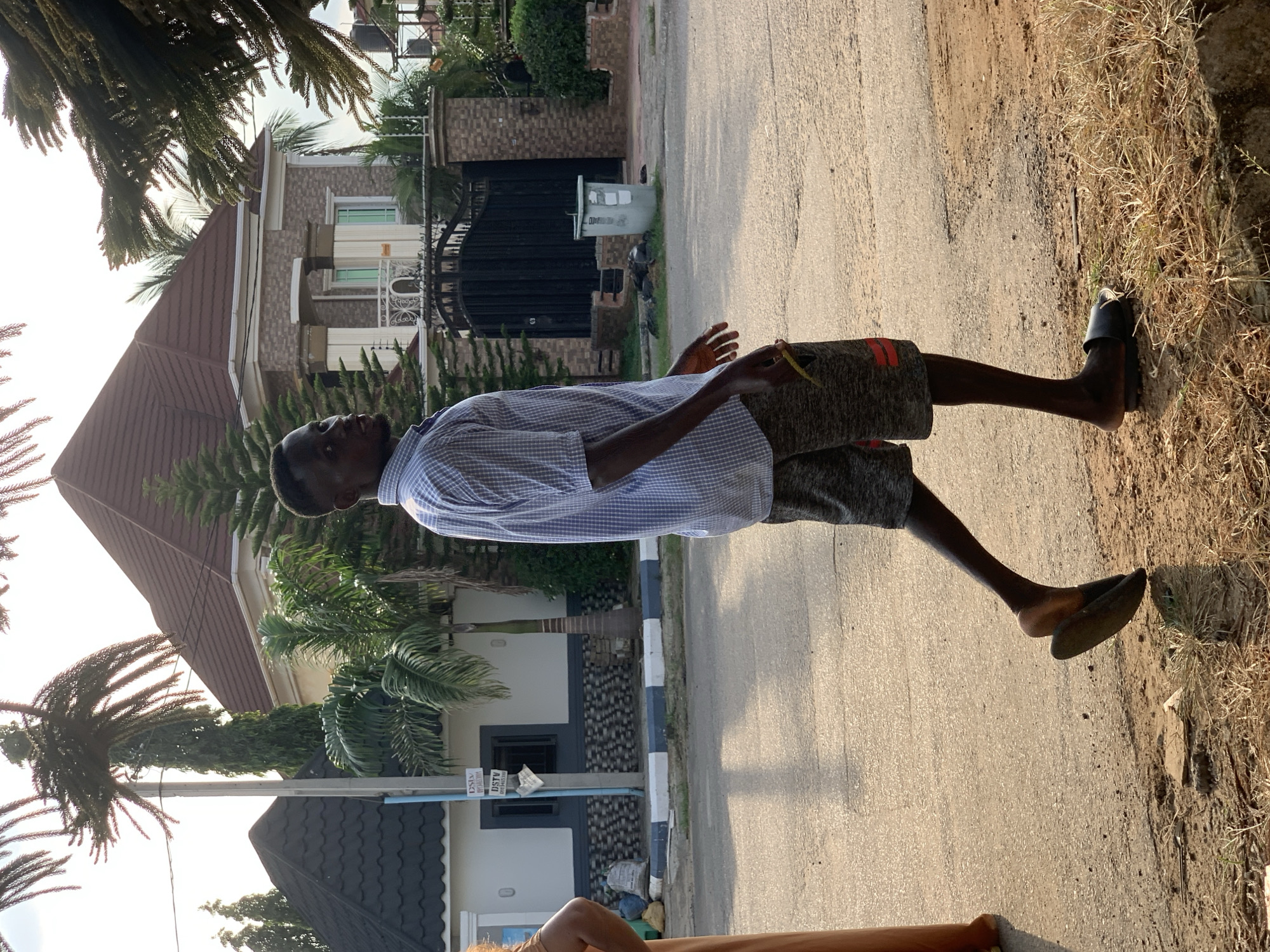 In the city of Abuja, Nigeria, a young Black man is captured in a full-body, medium shot, walking on a paved street, looking over his left shoulder towards the camera. He is wearing a blue and white checkered short-sleeved shirt and grey patterned shorts with red stripes on the sides. He has on black slide sandals. To his left, a person in a tan dress is partially visible. The background features a multi-story residential building with a brown, gabled roof and beige and grey exterior walls, characterized by columns and decorative railings. A large black wrought-iron gate is visible in front of the house. The street is lined with a curb painted with white and blue stripes. Palm trees and other dense green foliage are present throughout the scene, and the sky is overcast.