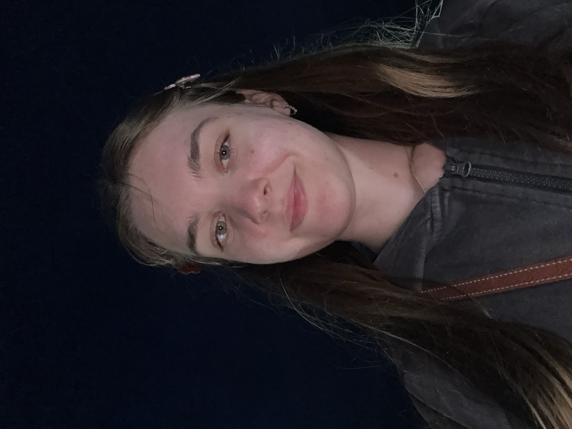 A young woman with light brown hair and light-colored eyes is photographed from the chest up. She is wearing a dark gray hooded jacket with a zipper that is partially open. She has a red hair clip on the left side of her head and a small hoop earring in her right ear. Her hair is long and falls around her shoulders. The background is a dark, solid color, likely indicating nighttime. No specific location in the city of Nizhniye Mulli, Russia, can be determined from the image.