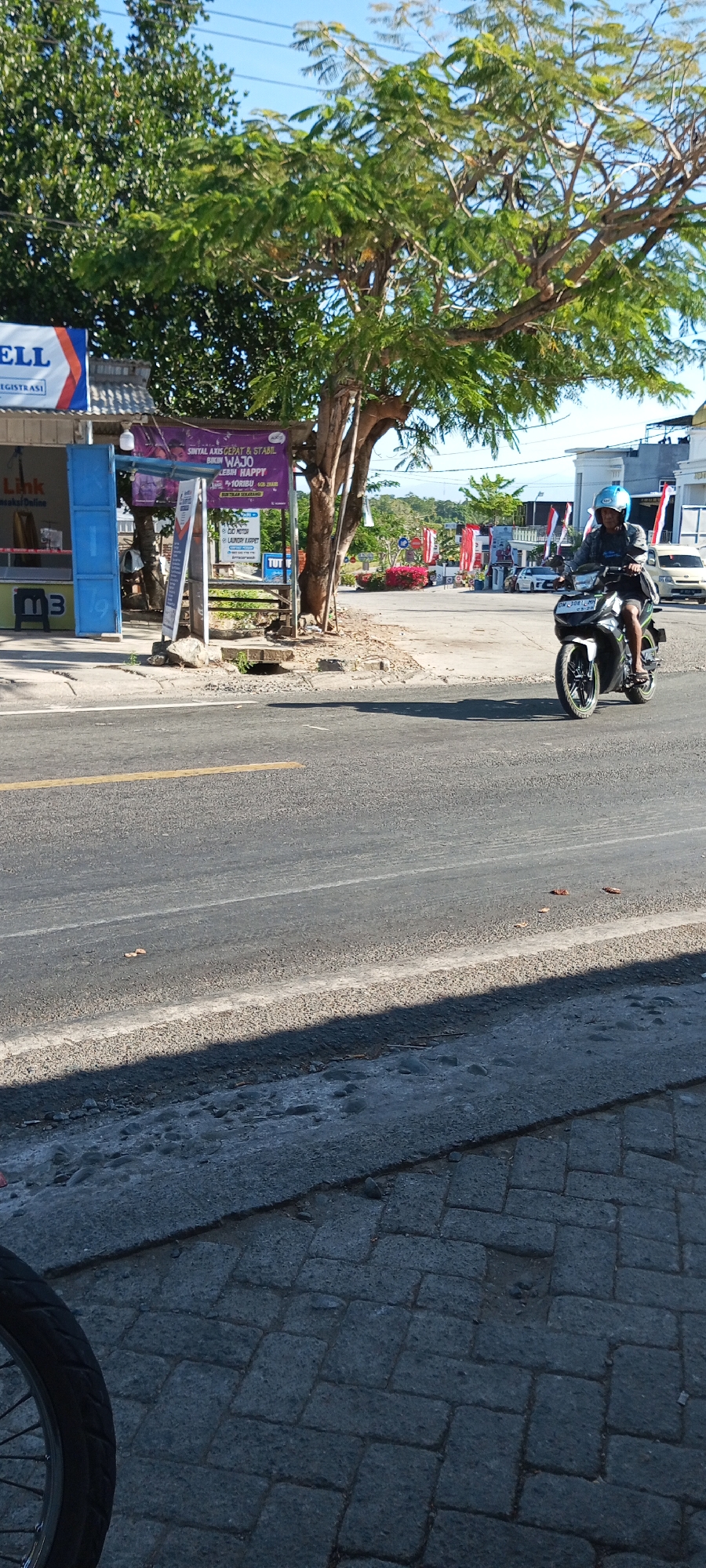 In the city of Pakkanna, Indonesia, a man wearing a blue helmet rides a black and green motorcycle on a paved road under a clear blue sky. The man is seen from the chest down, looking ahead as he rides. The motorcycle has a license plate visible. To the left of the road is a building with blue shutters and a sign that reads "Link". Further down the road, there are trees, a large tree with green leaves dominating the center of the frame. In the background, there are more buildings, some adorned with Indonesian flags. The road itself is grey asphalt with a yellow line marking the edge of a lane. A shadow from a nearby structure falls across the lower part of the image. To the far left, a portion of a bicycle wheel is visible.
