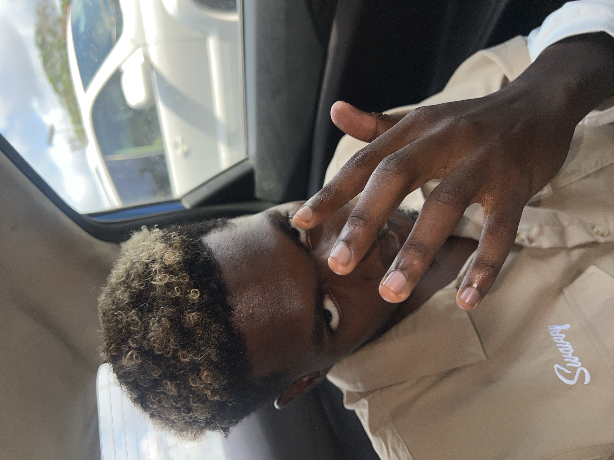 A close-up shot taken inside a vehicle shows a dark-skinned person with curly hair, wearing a light-colored shirt with "Swavy" embroidered on the chest, pressing their fingers against their eye. The backdrop reveals a view through the car window, suggesting the setting may be an urban environment in Lagos, Nigeria, though no distinct Lagos landmarks are visible. The lighting indicates daytime.