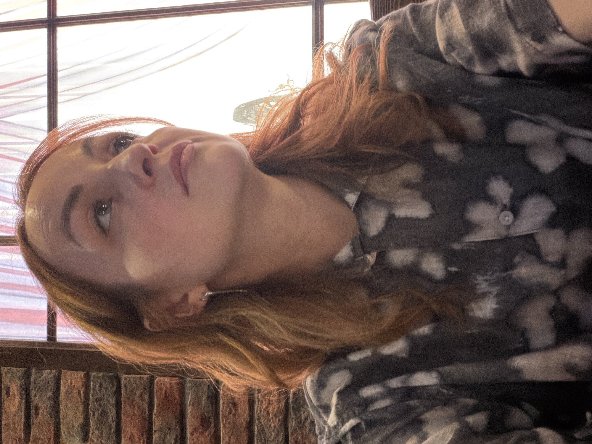 A medium shot depicts a woman indoors. Her head is tilted upward and slightly to the left. She has fair skin and light brown hair, which appears to be highlighted with red tones. She is wearing an earring in her left ear and a printed, button-up shirt. The background shows a window with visible window panes and a blurry, bright outdoor scene. Below the window is a brick wall. The location is not discernible from the image alone.