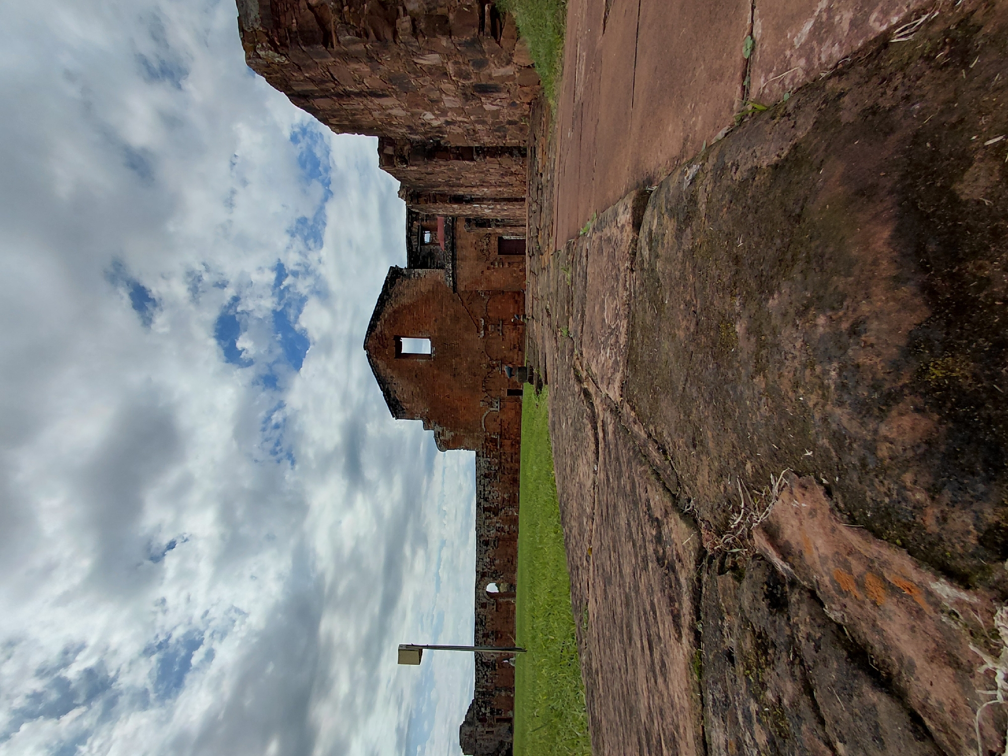 An outdoor, eye-level, wide-angle photograph captures the ruins of the Santísima Trinidad de Paraná mission, a UNESCO World Heritage site located in the city of Trinidad, Paraguay. The foreground features large, weathered, reddish-brown stone pavers, some cracked and covered in moss, leading the eye towards the dilapidated brick structures of the former Jesuit mission. These ruins are primarily made of red brick, with some walls and archways still standing, though many have collapsed, revealing empty window frames and crumbling interiors. The sky above is filled with dramatic, grey and white clouds, interspersed with patches of blue sky. To the left, a manicured lawn of vibrant green grass stretches towards a long, low brick wall, punctuated by an arched opening. A single, thin, grey pole with a light-colored rectangular fixture stands on the lawn, near the wall. The overall impression is one of historical grandeur and decay under a dynamic sky.