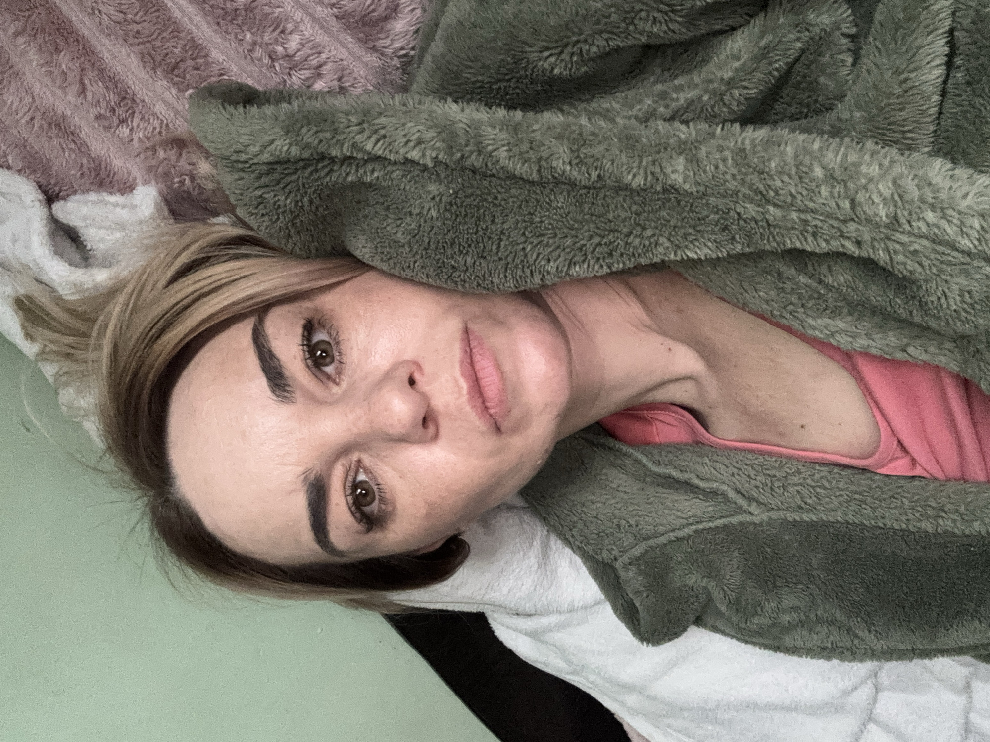 A woman is lying down. She is partially covered by a green, furry blanket. She is wearing a pink top. A white towel and a pink blanket are also visible. The location is Unknown, Russia.