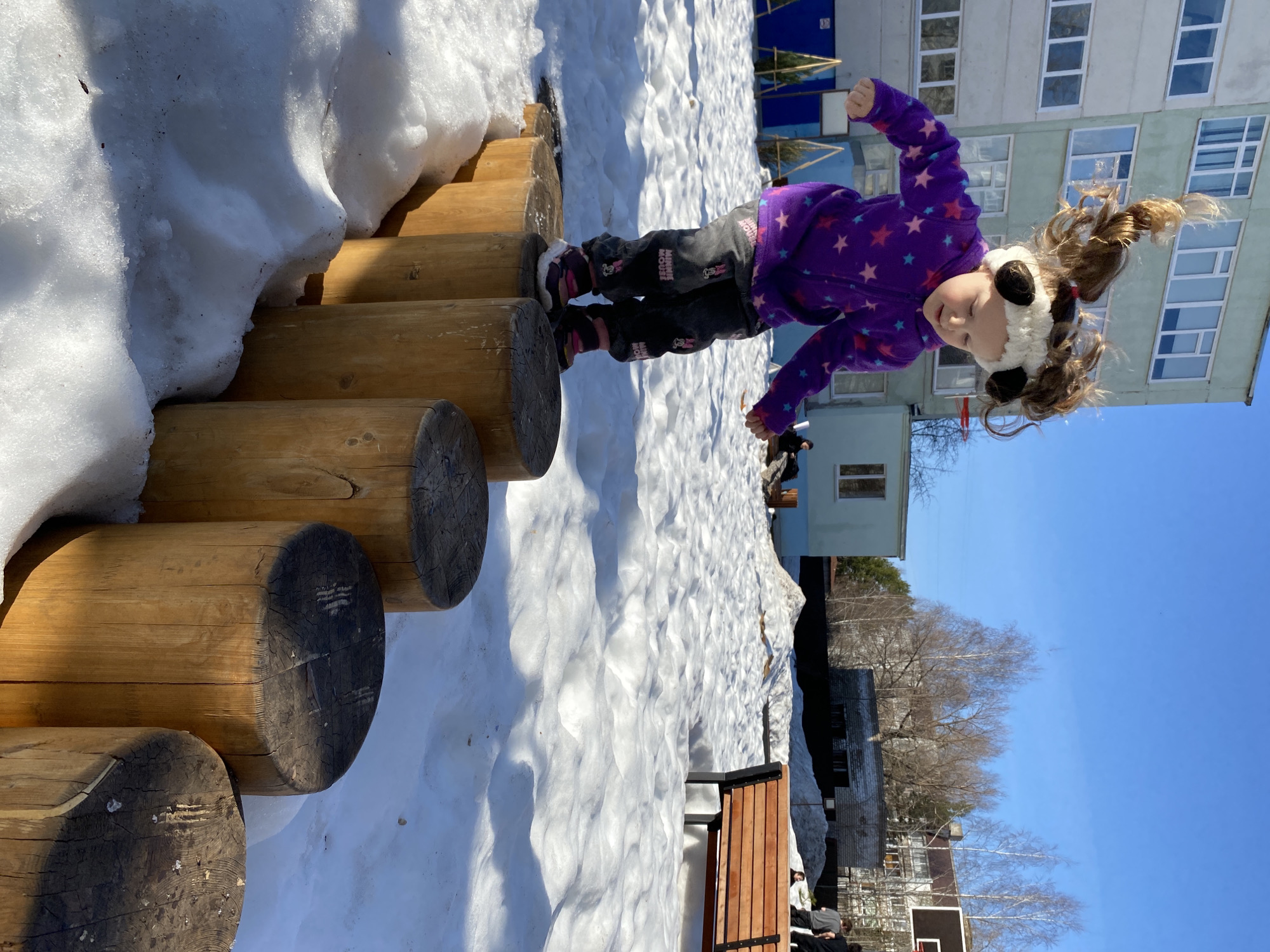 A young girl in a purple star-patterned jacket and panda-ear hat is standing on a wooden log structure, partially covered in snow, in Chaikovsky, Russia. The background shows a light-colored building and a clear blue sky. A snow-covered ground surrounds the structure.