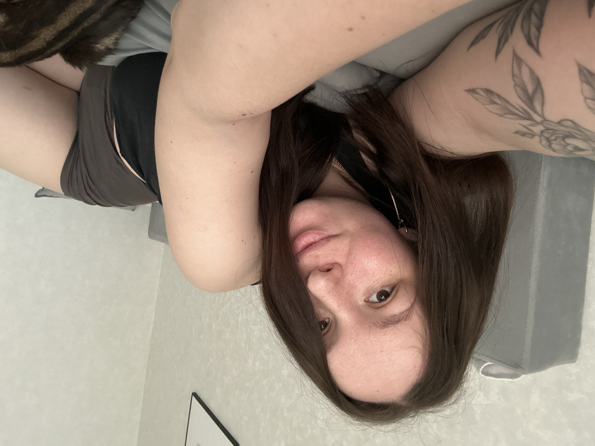 A female individual is depicted in a close-up selfie, lying partially on her side in an indoor setting located in Chaikovsky, Russia. She has fair skin, dark brown hair, brown eyes, and visible freckles on her face. A floral tattoo is present on her left upper arm. She is wearing a dark-colored top and dark brown shorts. Long, chain-style earrings are visible. Her right arm is extended, indicating she is holding the device capturing the image. She looks directly at the camera with a slight smile. Behind her left shoulder is a grey upholstered headboard or cushion, and the wall features light-colored, textured wallpaper with a minimalist black-framed picture in the upper right. Grey bedding is visible beneath her. A brown tabby cat, partially visible in the bottom right, appears to be resting near the woman.