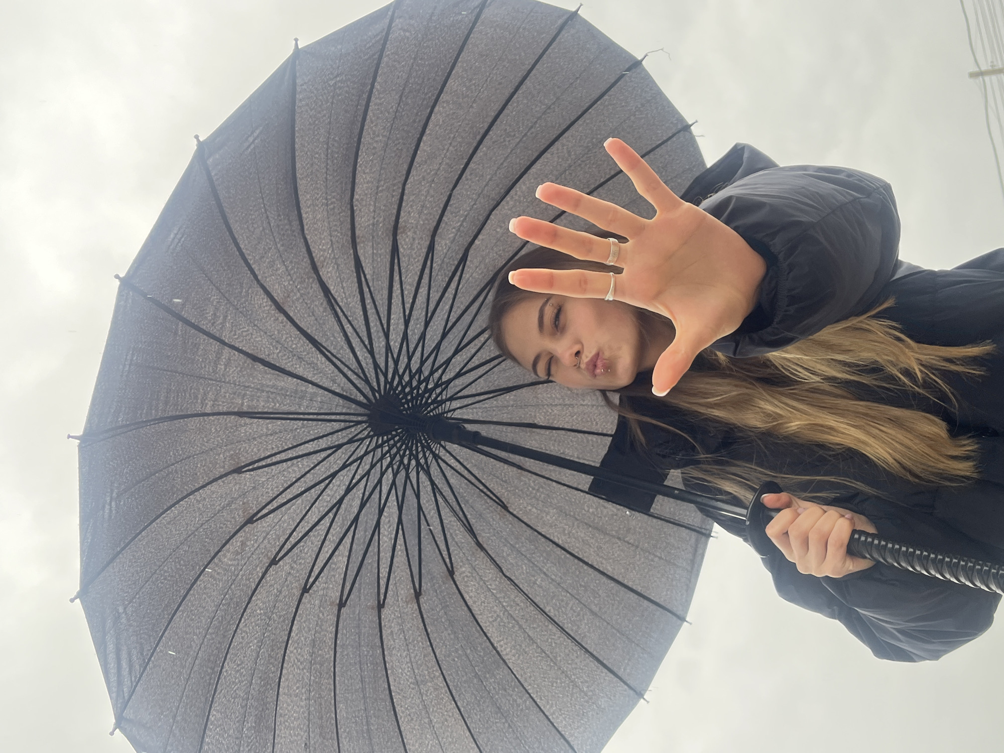A low-angle shot captures a person holding a large, dark grey umbrella against a cloudy sky. The person is wearing a dark jacket. Their blonde hair is visible along the right side of the jacket. The person is making a kissing face, and their right hand is outstretched towards the camera. The hand has two rings on the fingers. The location is Фоки, Russia.