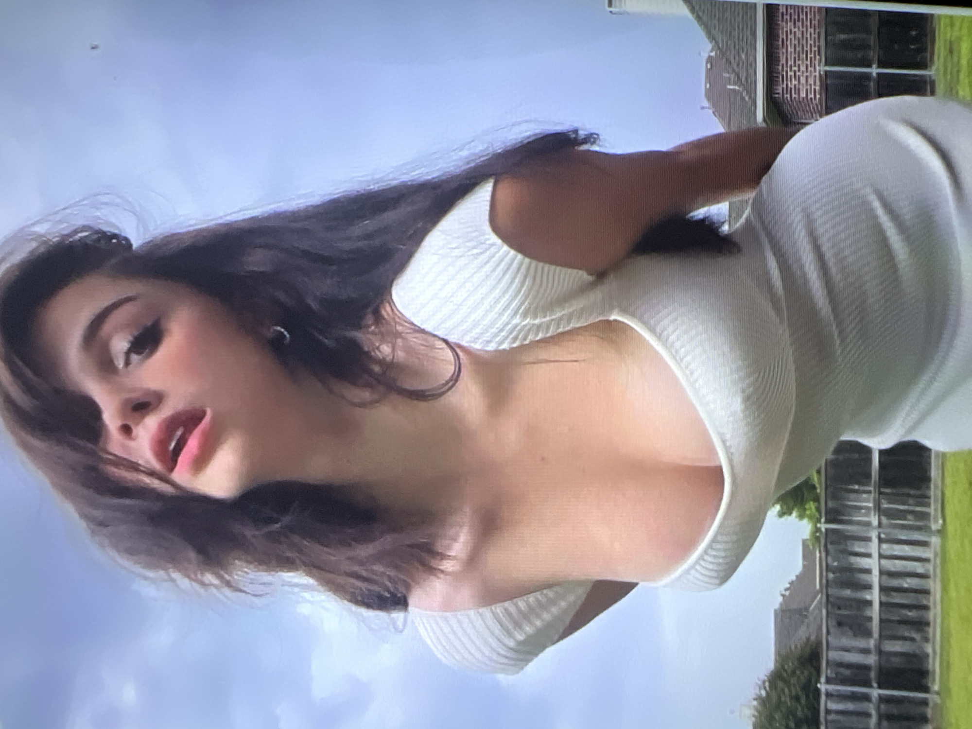A young woman with dark hair and fair skin is photographed outdoors. She is wearing a white, ribbed, short-sleeved dress that accentuates her figure. The background shows a wooden fence, green grass, and residential buildings under a cloudy sky. The woman's gaze is directed upwards and away from the camera, with her mouth slightly open. Her makeup is visible, with emphasis on her eyes and lips. The image appears to be a candid shot, possibly taken from a low angle. There is no discernible information in the image to suggest the location of Energetik, Russia.