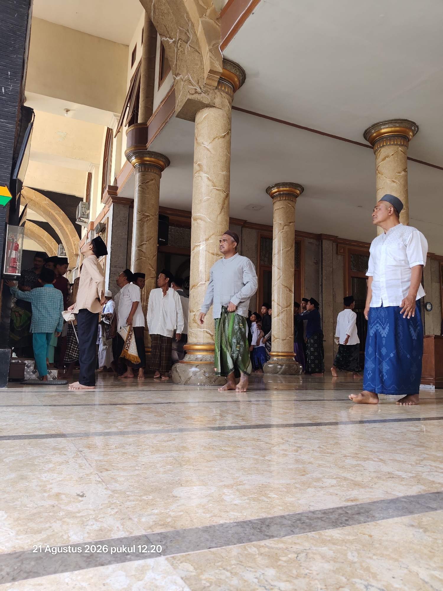 The image depicts a group of people, predominantly men and boys, gathered in an indoor space with large marble-patterned floors and ornate columns supporting an arched ceiling. The architecture suggests a public building, possibly a mosque or a community hall, in Kalitidu, Indonesia. Most of the individuals are wearing traditional Indonesian attire, including sarongs and peci caps. Some are barefoot. In the foreground, two men are standing, one in a white shirt and blue sarong, and another in a grey shirt and green sarong. To the left, a young boy in a plaid shirt and blue pants is looking up at a display case on a dark wall. The overall atmosphere appears to be one of a gathering or a moment of pause. The timestamp "21 Agustus 2026 pukul 12.20" is visible at the bottom left of the image, indicating the date and time the photograph was taken.