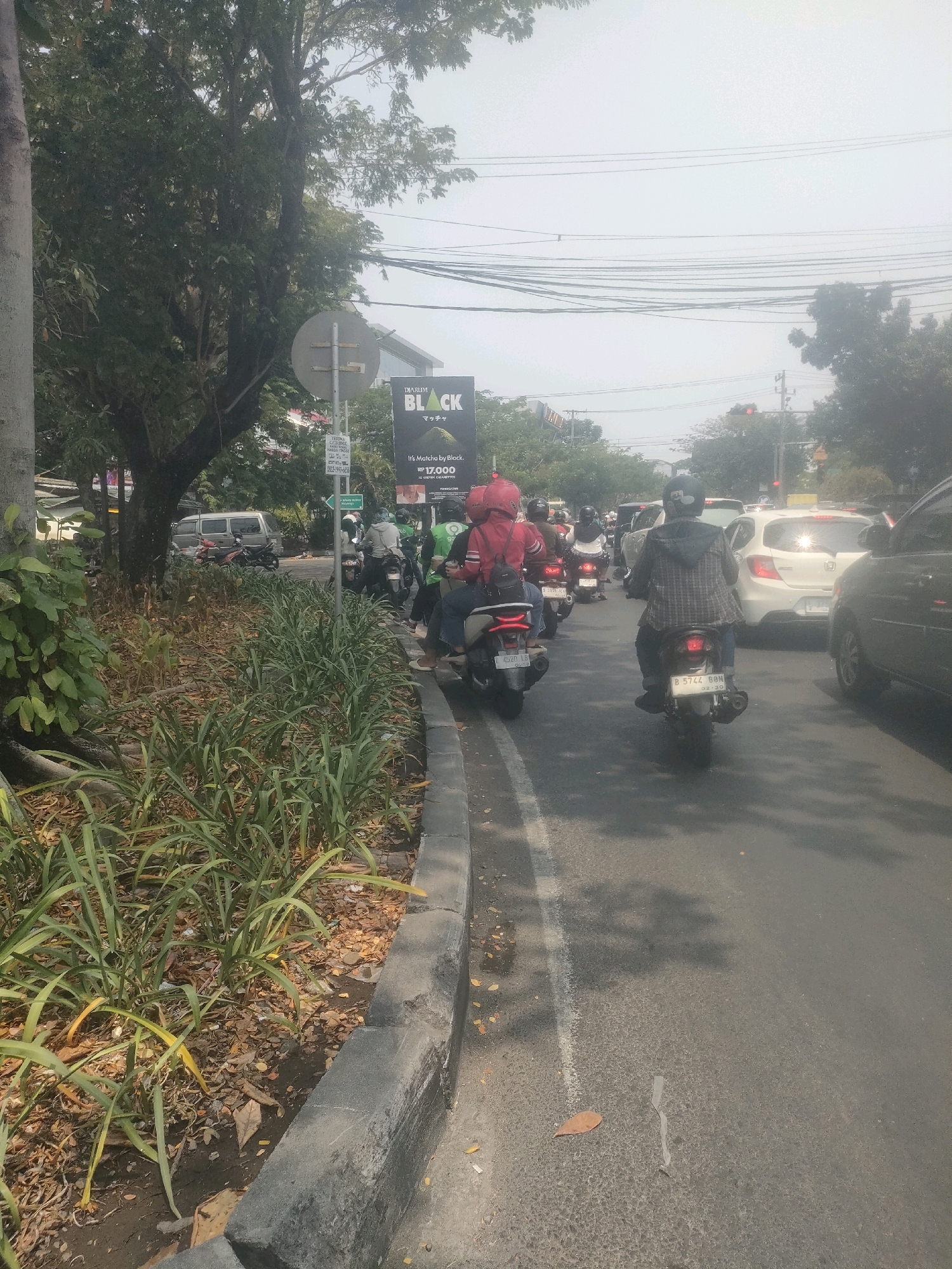 A daytime, eye-level outdoor shot captures a busy street scene in Surabaya, Indonesia, with a focus on traffic and urban elements. The foreground features a concrete curb bordering a patch of overgrown green plants and dry, fallen leaves. The street itself is paved and marked with a white line, with a slight sheen indicating recent moisture or a slick surface.
The middle ground is dominated by a line of motorcycles and a few cars, indicating moderate traffic congestion. Several individuals on motorcycles are visible, most wearing helmets, suggesting adherence to safety regulations. Some riders are wearing casual clothing, while others, possibly delivery personnel, wear uniforms. The motorcycles range in color and design, reflecting common models in the region. A white car with its brake lights illuminated is also present in the traffic.
In the background, lush green trees line the street, interspersed with buildings and overhead power lines. A large billboard advertising "DIARUM BLACK" with "It's Matcha by Black." and "RP 17.000" is prominently displayed, suggesting commercial activity. The sky is hazy and bright, typical of a sunny day. The overall impression is a snapshot of daily urban life in Surabaya, characterized by bustling traffic and the presence of natural elements within the city.