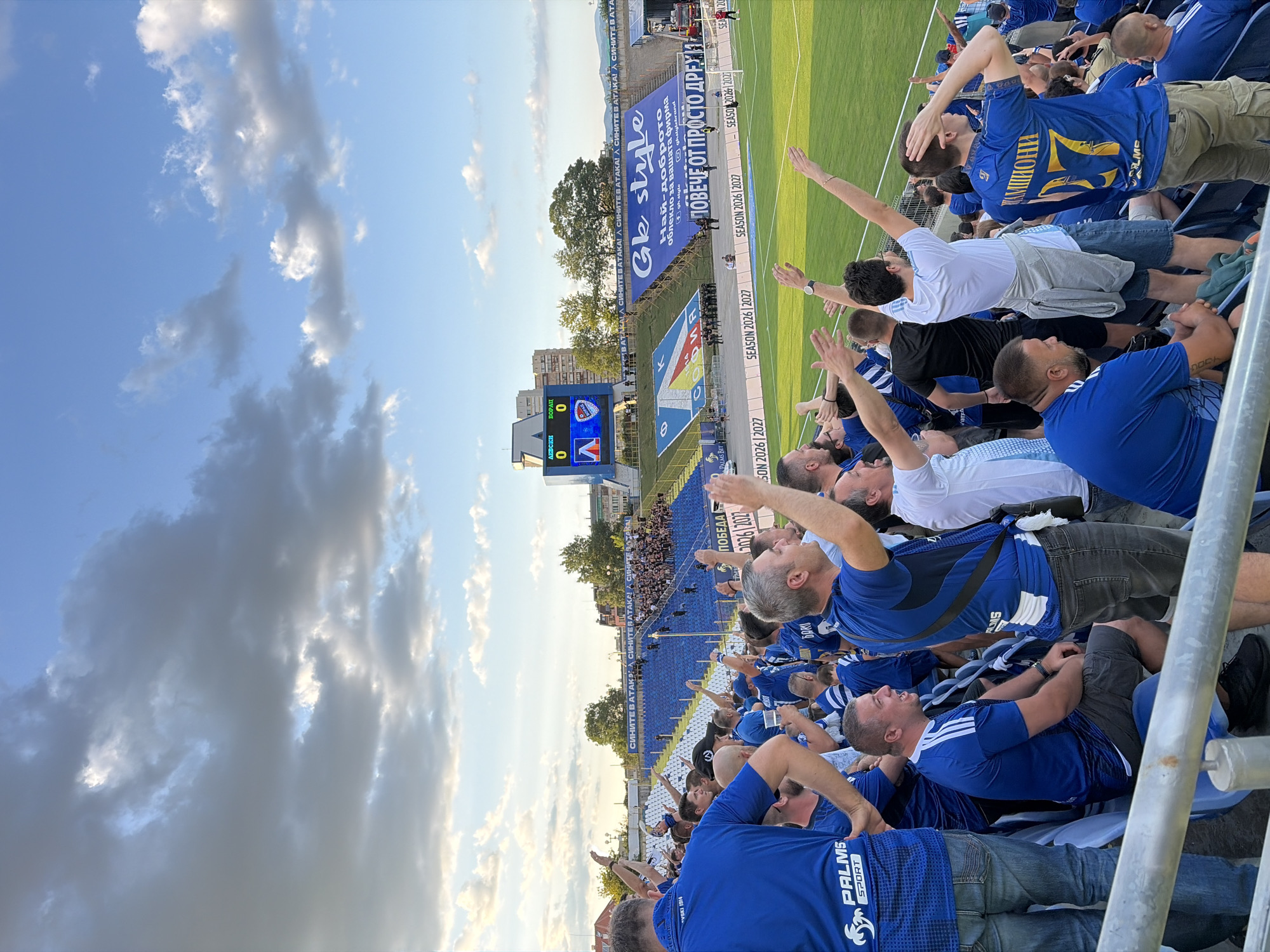 This is a medium shot of a crowd of football fans cheering in a stadium. The fans are wearing blue jerseys and are positioned in the stands, with the football field visible in the background. Some fans are raising their hands in the air, and others are holding drinks. There is a scoreboard in the distance that shows "0-0". There is a large banner in the background that reads "SEASON 2026 2027". The stadium appears to be located in Sofia, Bulgaria, as indicated by the text on the scoreboard which reads "АСКИ 0" and "БОРАК 0" and other visible Bulgarian text on banners and jerseys. The sky is partly cloudy.