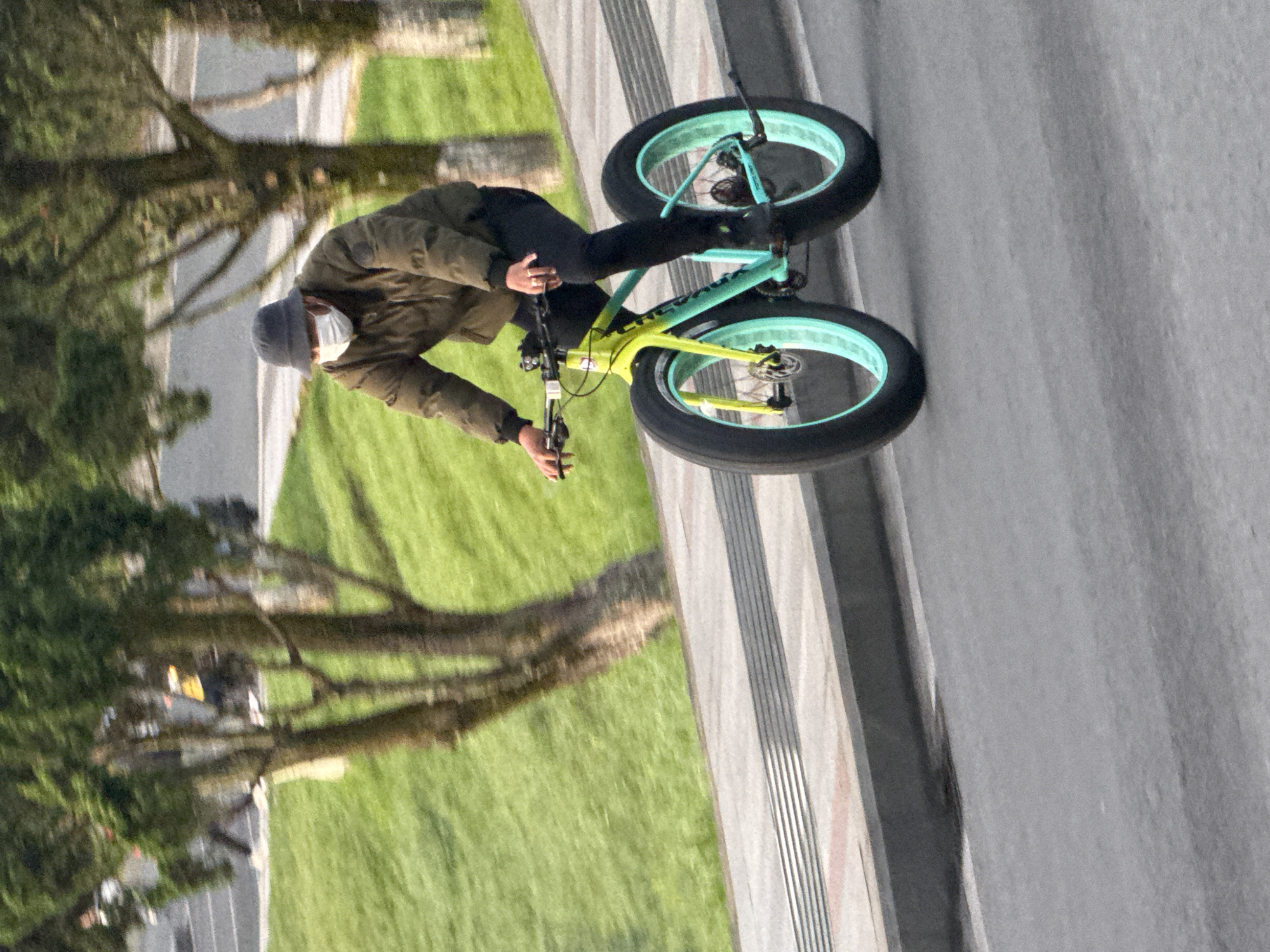 In Da Lat, Vietnam, a person wearing a dark bucket hat and a white face mask rides a bright yellow and teal fat-tire bicycle. They are dressed in a dark olive green puffer jacket and black pants, with black shoes. The rider is on a paved road, with a textured grey and red sidewalk bordering a grassy slope to their left. Several trees with green foliage are visible in the background. The lighting suggests it is daytime, with shadows indicating the sun is at a medium angle. The image is a medium shot focused on the cyclist and their bicycle.