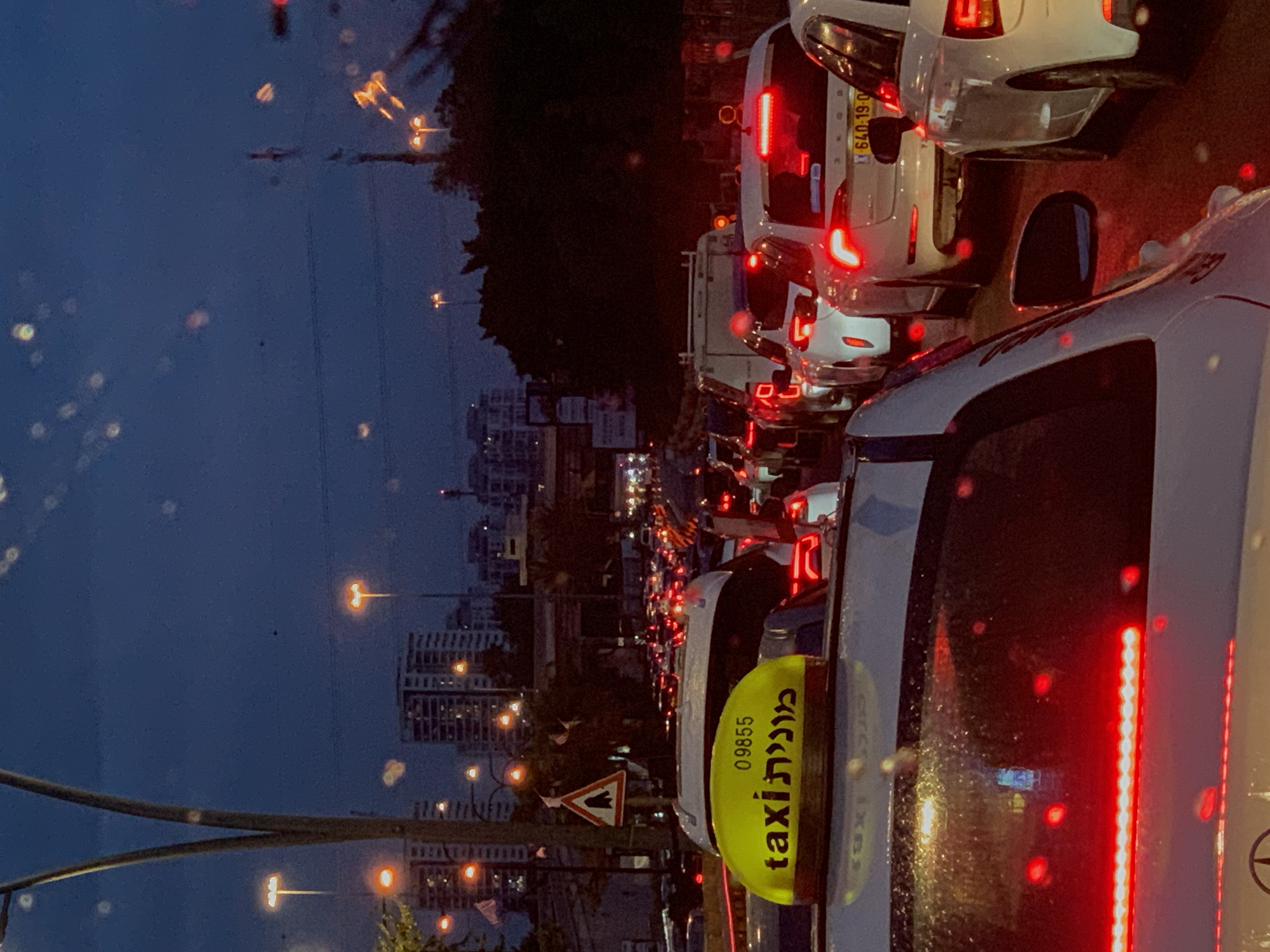 The image depicts heavy traffic congestion in Ramat Gan, Israel, at dusk or night. A line of cars with illuminated taillights stretches into the distance, suggesting a significant traffic delay. A taxi with a visible yellow roof sign displaying "taxi" in both Hebrew and English is prominently featured in the foreground. Streetlights and buildings are visible in the background, indicating an urban environment. Raindrops or condensation are present on the window, blurring the view slightly. The overall scene conveys a sense of congestion and urban activity.