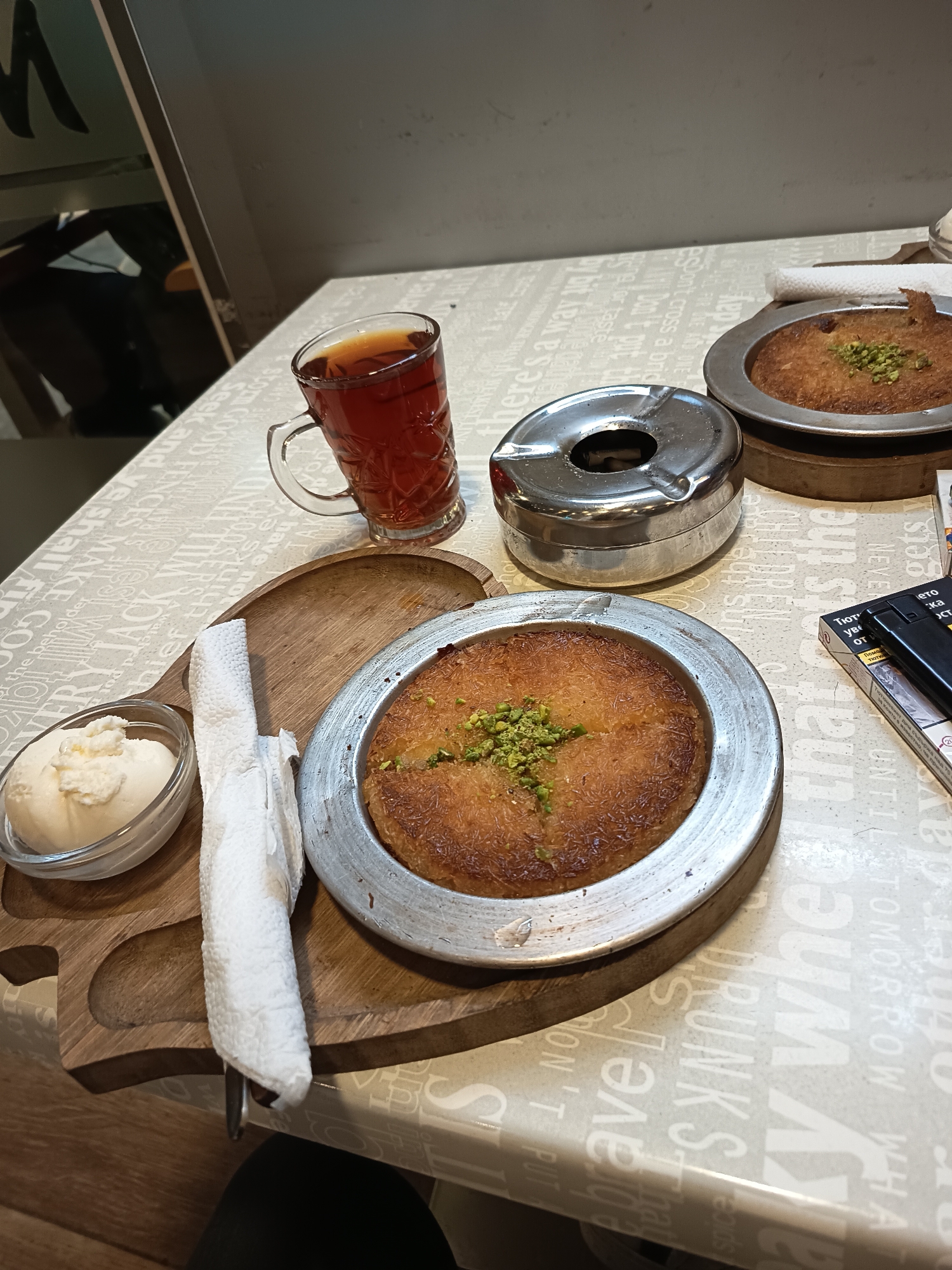 The image depicts a table setting, likely in a café or restaurant in Sofia, Bulgaria, judging by the Cyrillic on the cigarette package. The tabletop has a patterned surface with English words and phrases printed on it.
There is a clear glass mug of dark tea, a metal ashtray, and two plates of what appears to be "künefe," a Middle Eastern cheese pastry soaked in sweet, sugar-based syrup. One plate is served with a side of white cream, perhaps kaymak or a similar dairy product, and a neatly folded white napkin.
A wooden serving tray holds one of the künefe plates along with the cream and napkin. A package of cigarettes and a lighter are also visible on the table. No people are discernible in the immediate scene, suggesting the photo was taken before or after a customer interaction. The environment appears indoors with a partial view of a chair and possibly a window or mirrored surface in the background.