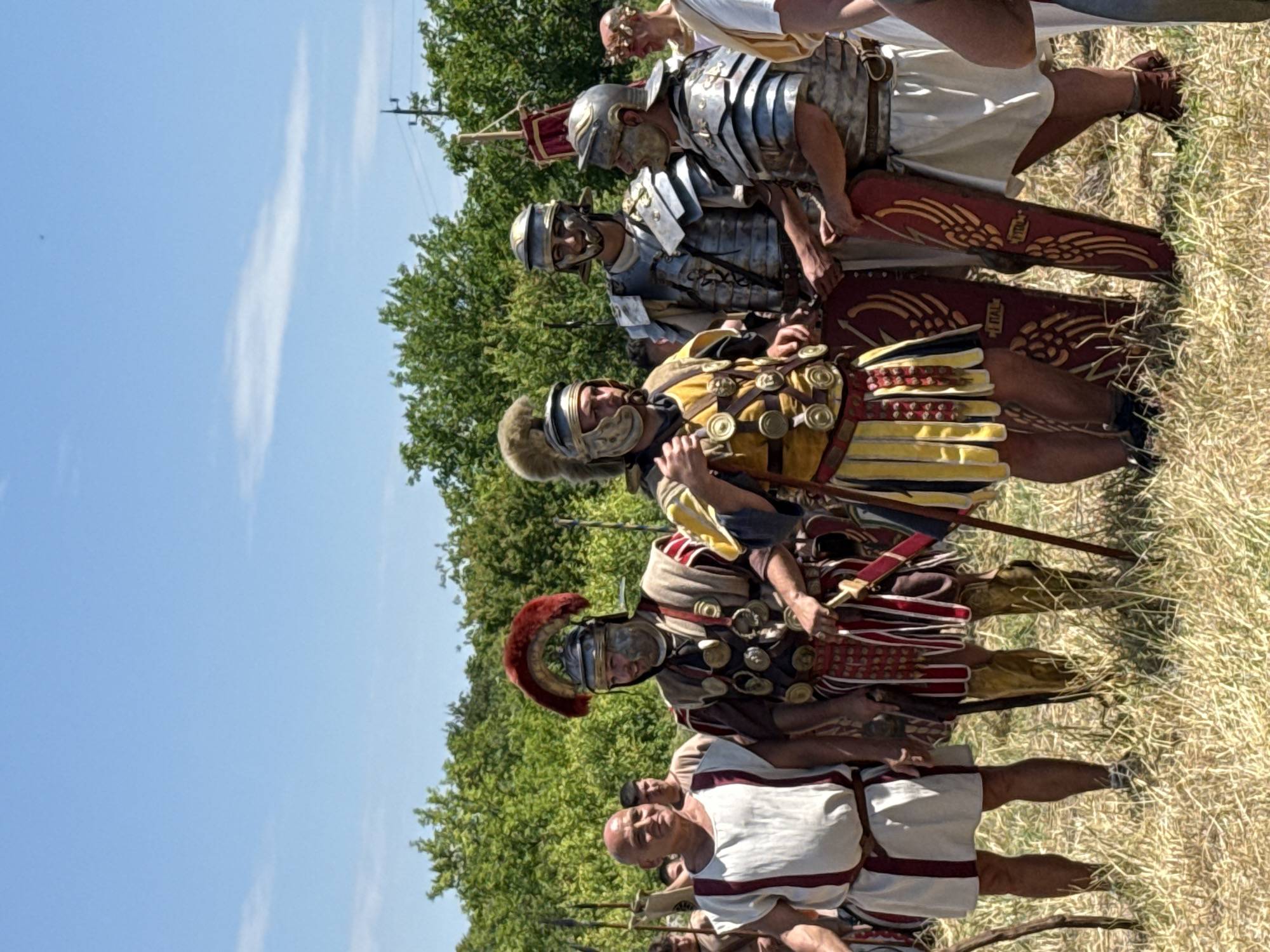 The image depicts a historical re-enactment scene in Gigen, Bulgaria, featuring a group of individuals dressed in ancient Roman attire standing outdoors on a sunny day. The foreground consists of dry, light-colored grass and sparse vegetation, transitioning to dense green deciduous trees in the midground and background. The sky above is clear blue with minimal scattered white clouds. A utility pole with wires is visible against the tree line in the upper right quadrant.
Six prominent individuals are visible in the central frame, primarily dressed as Roman legionaries. From left to right:
1. A bald man in a white tunic with maroon vertical stripes, smiling and looking towards the center-right. He holds a wooden staff.
2. A legionary wearing a helmet with a prominent red plumed crest, segmented body armor (lorica segmentata) over a dark tunic, and multiple large circular medallions on his chest piece. He is looking right, armed with a gladius at his left hip.
3. A legionary in a helmet with a fur-like crest and a gold-striped tunic over body armor adorned with medallions. He is holding a red scutum (shield) with gold wing-like designs, bearing the partial inscription "ITAL".
4. A legionary in silver-toned segmented armor and helmet, holding a red scutum with gold wing-like designs, also bearing a partial "ITAL" inscription. He is turned towards the right, appearing to interact with the next figure.
5. Another legionary, similarly attired in silver-toned segmented armor and helmet, facing left and engaged in conversation with the previous figure. He also holds a red scutum with gold wing-like motifs.
6. A partially visible figure to the far right, wearing a white tunic and a golden wreath on his head, possibly a civilian or dignitary.
Additional figures in Roman period clothing are visible in the background, standing among the trees. The overall scene suggests an organized historical re-enactment or festival under bright daylight conditions.