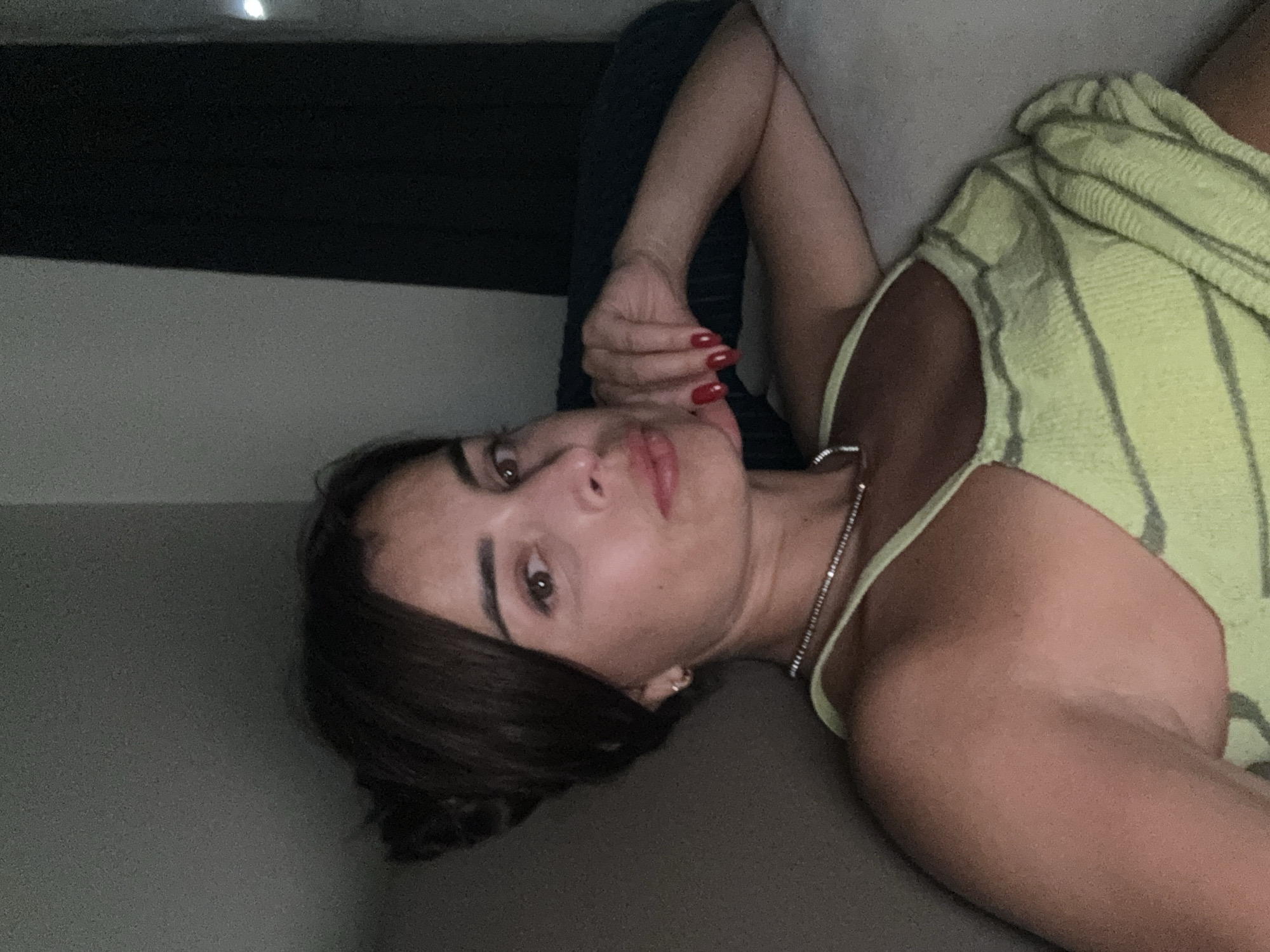 An overhead shot shows a woman with dark hair pulled back, wearing a lime green striped top and a delicate silver necklace. Her right arm is bent, with her hand resting on her cheek, and her fingernails are painted red. She is reclining on a light-colored surface, possibly a couch or bed, with a dark background behind her. The lighting is dim, creating a shadowed environment.