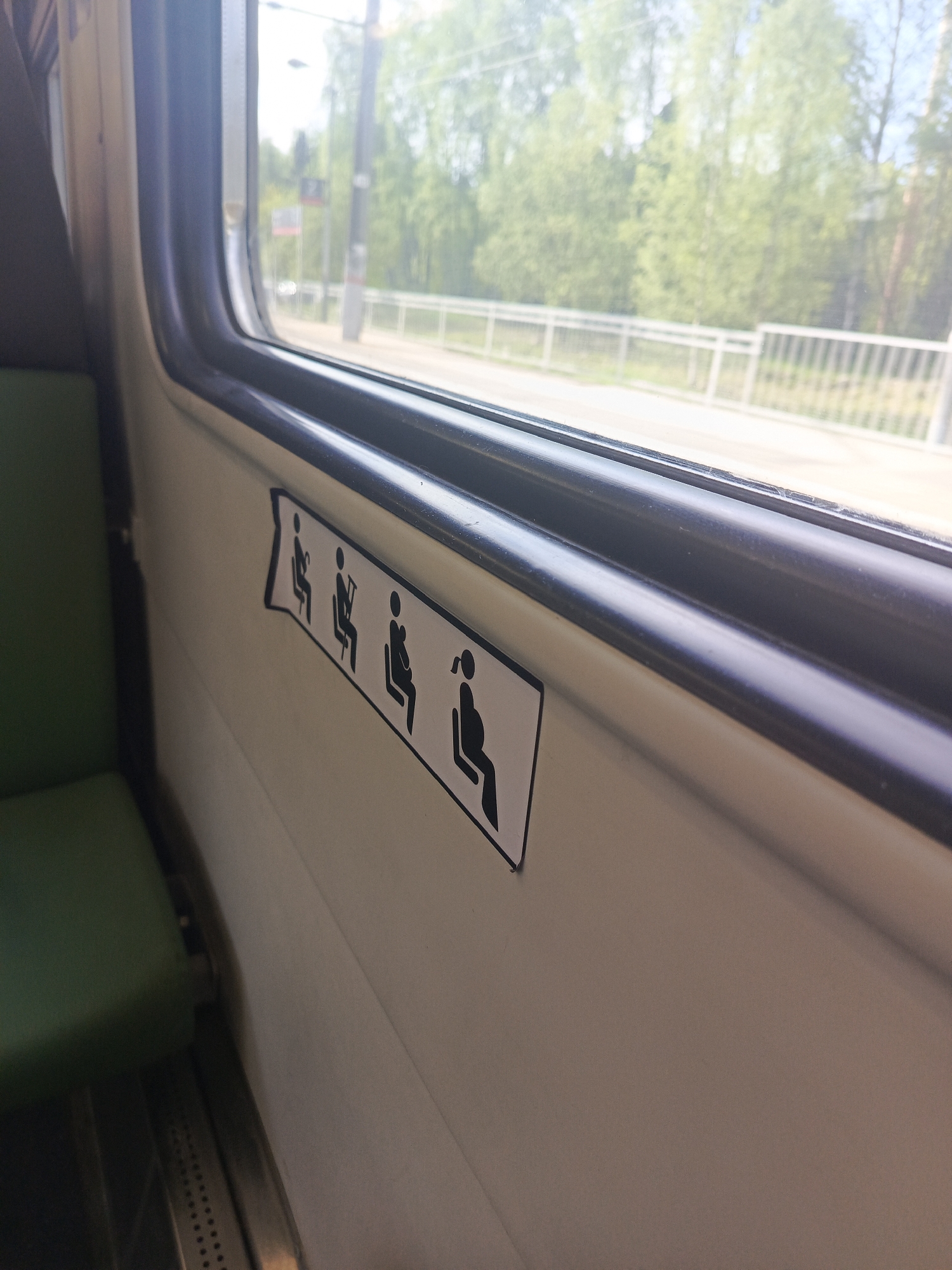 The interior of a train car is shown. A green-cushioned seat is visible to the left. Adjacent to the seat is the train car wall, upon which is affixed a sign indicating priority seating. The sign shows, in sequence, seated silhouettes of an able person, an elderly person with a cane, a person with a cast on their leg, and a pregnant person. Outside the window is a view of a tree-lined roadway with a bridge. The location is Unknown, Russia.