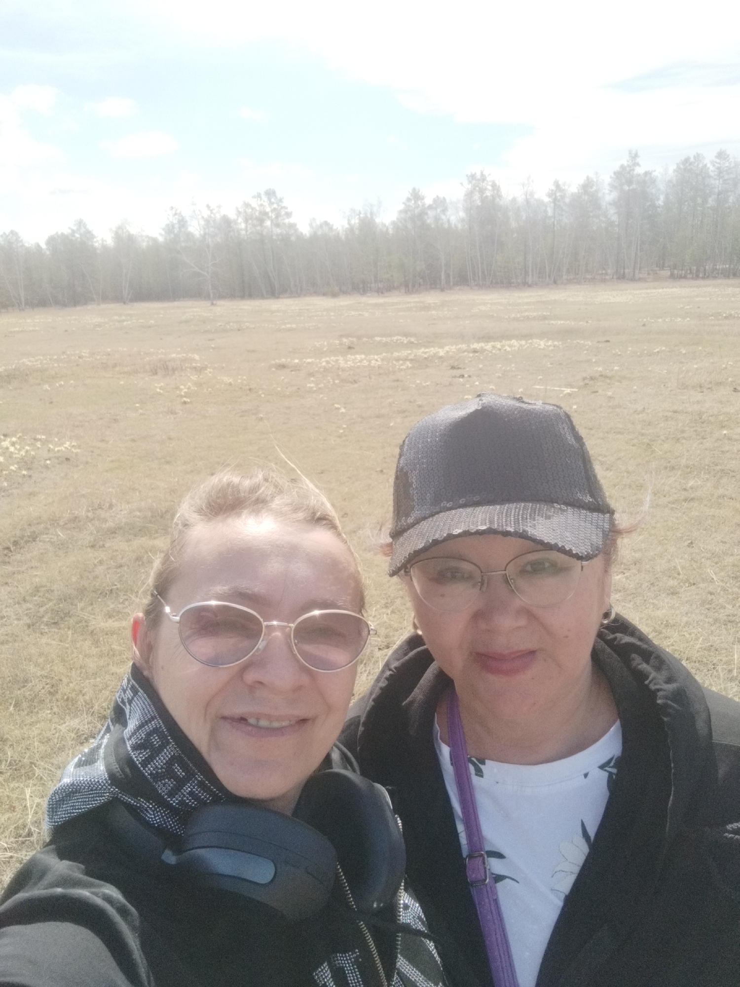 Two women are standing close together, facing the camera, in an open field. Behind them is a field of grass and small white flowers, and in the background, there's a line of trees. The woman on the left has blonde hair and is wearing round sunglasses and a black jacket with a patterned scarf. Headphones are around her neck. The woman on the right is wearing glasses and a sequined baseball cap, a black jacket, and a white floral-patterned shirt. A purple strap is visible across her chest. The sky is bright. The location is Syrdakh, Russia, as specified.