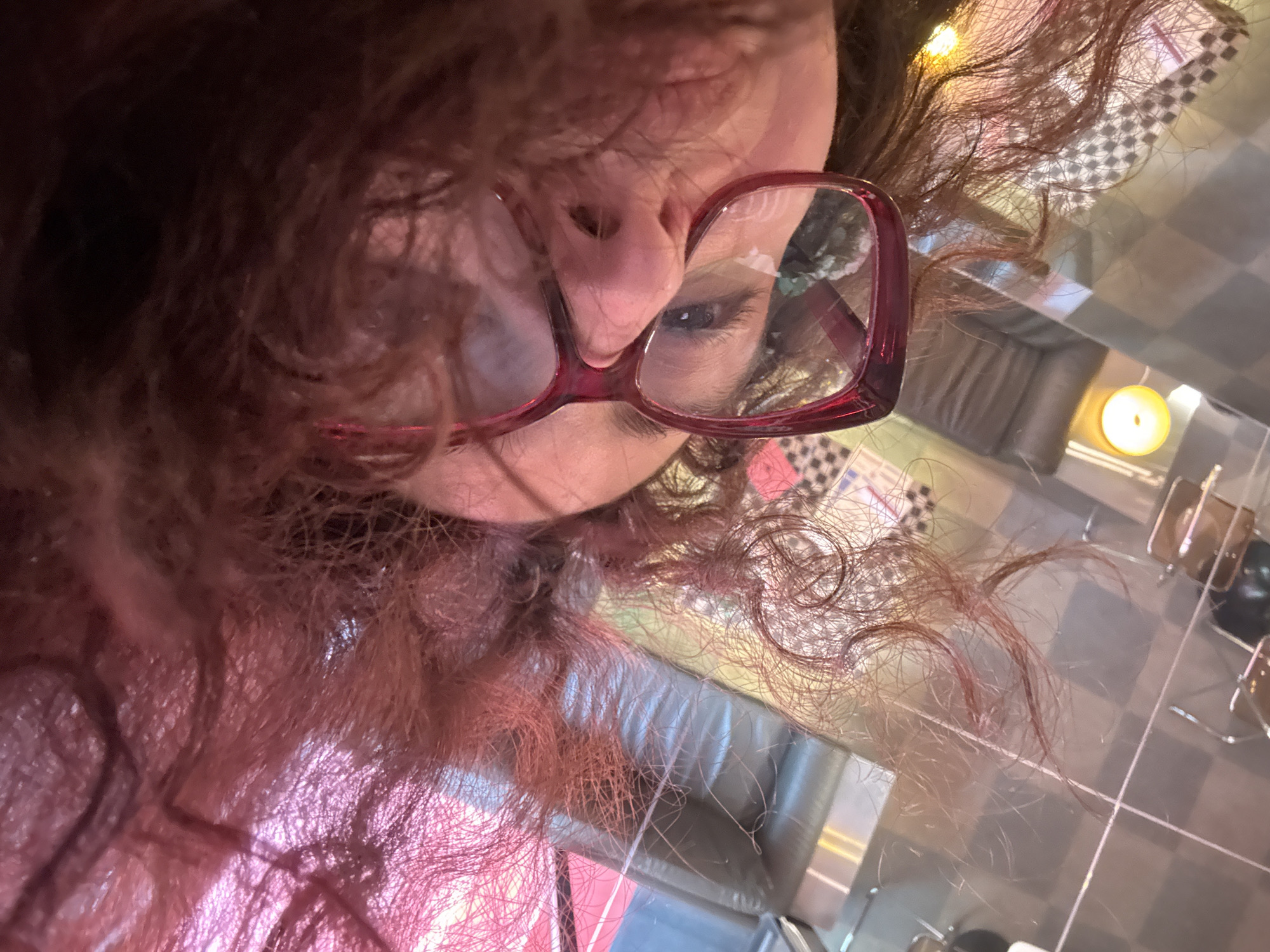 The image shows a close-up, upside-down view of a person with red curly hair and glasses. The person is looking at the camera. The background is blurry but appears to show a table with a checkered pattern, a candle, and furniture. The location is not identifiable as Yerevan, Armenia based on the provided image.