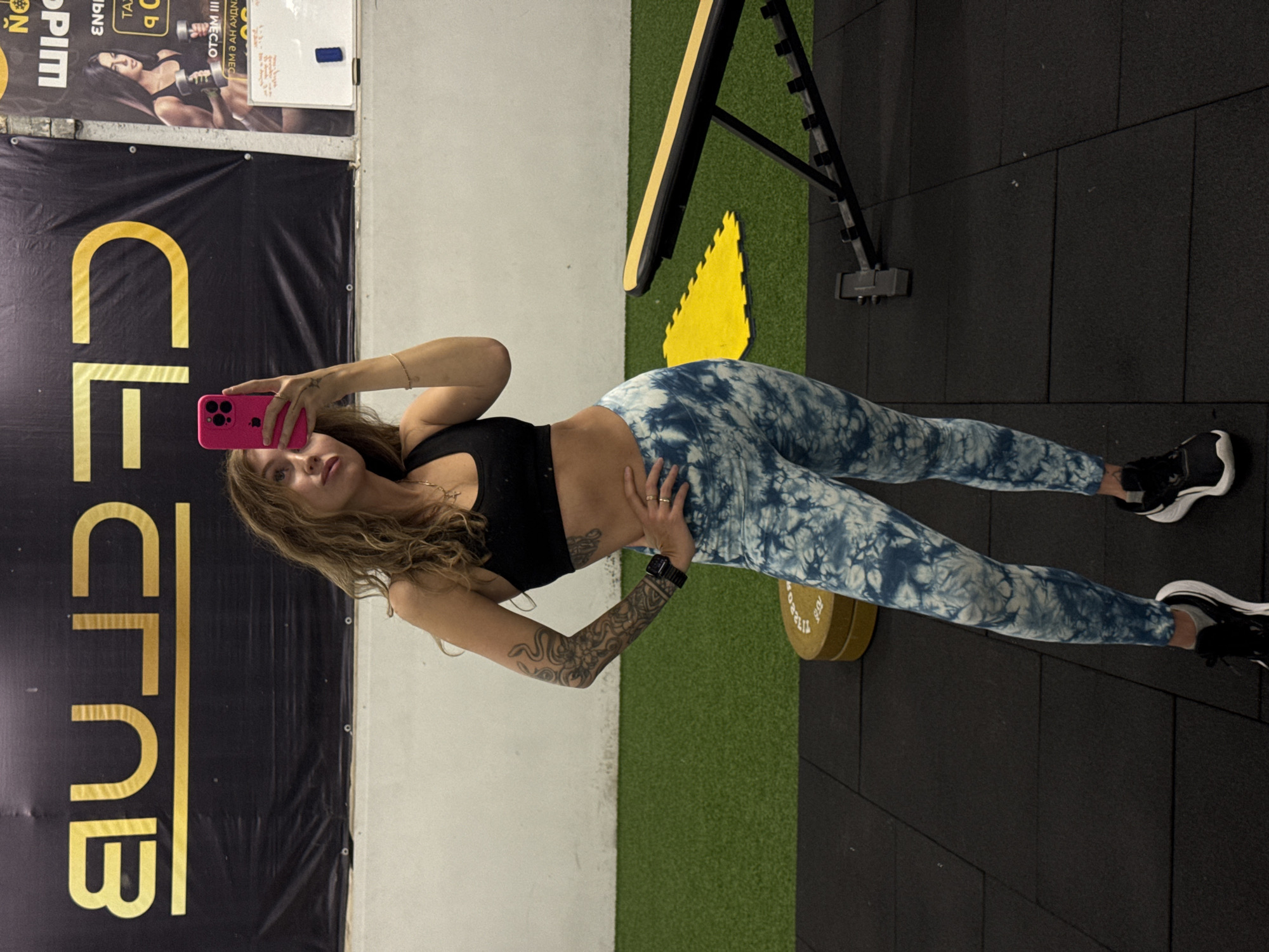 This media file captures a young woman posing for a photo inside a gym or fitness club, located in Chaikovsky, Russia. She stands upright, looking towards the camera, holding a bright pink smartphone in her right hand, likely taking a selfie. She has long, wavy, light brown hair and a neutral to subtly smiling expression. Her attire consists of a black sports bra that exposes her midriff and high-waisted blue and white tie-dye patterned leggings. She has a full sleeve tattoo on her left arm with intricate designs and wears a watch on her left wrist, along with rings on her right hand.
The background provides typical gym setting cues. To her left, a large black banner with bold gold lettering spells out "CLUB". Above this banner, another poster is partially visible, displaying fragments of text such as "NO PIM", "EN9B", and "TA", which appear to be in Russian, consistent with the location context. A whiteboard with some illegible handwritten notes is also present near the top. To her right, gym equipment is visible, including a black weight bench and a yellow circular weight plate, clearly marked "112.20 LBS" (approximately 50 kilograms) along with other numbers. The floor is a combination of black rubber tiles and green artificial turf. The walls are a plain light color, and the indoor lighting suggests it is daytime.